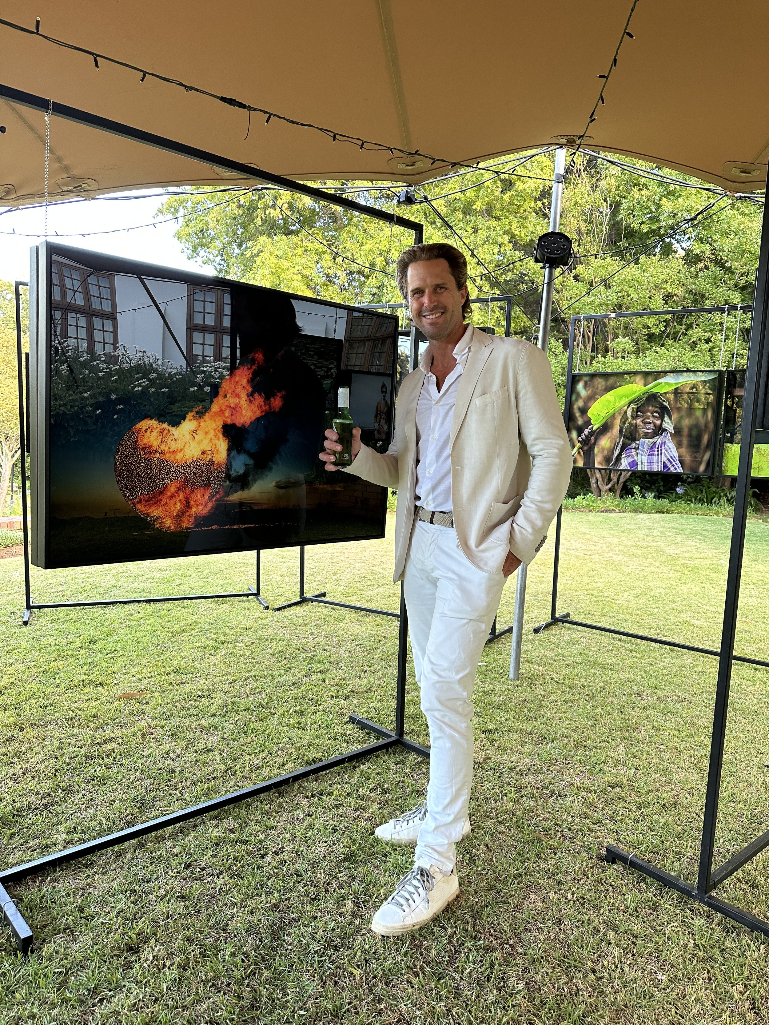 A man in a white suit and white sneakers smiling and holding a drink, standing outdoors under a tent with string lights, surrounded by display screens showing images of fire and a person with a green hat.