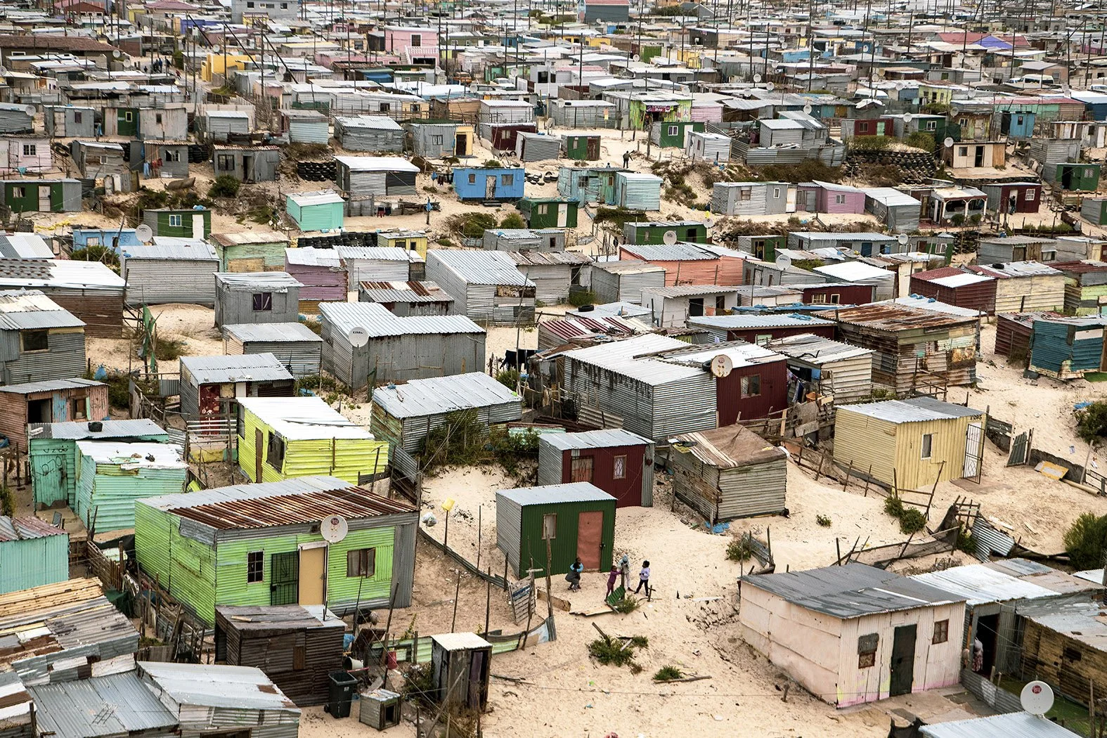 A densely populated informal settlement with makeshift homes made from corrugated metal and wood, set on sandy terrain, with a few people walking among the houses.