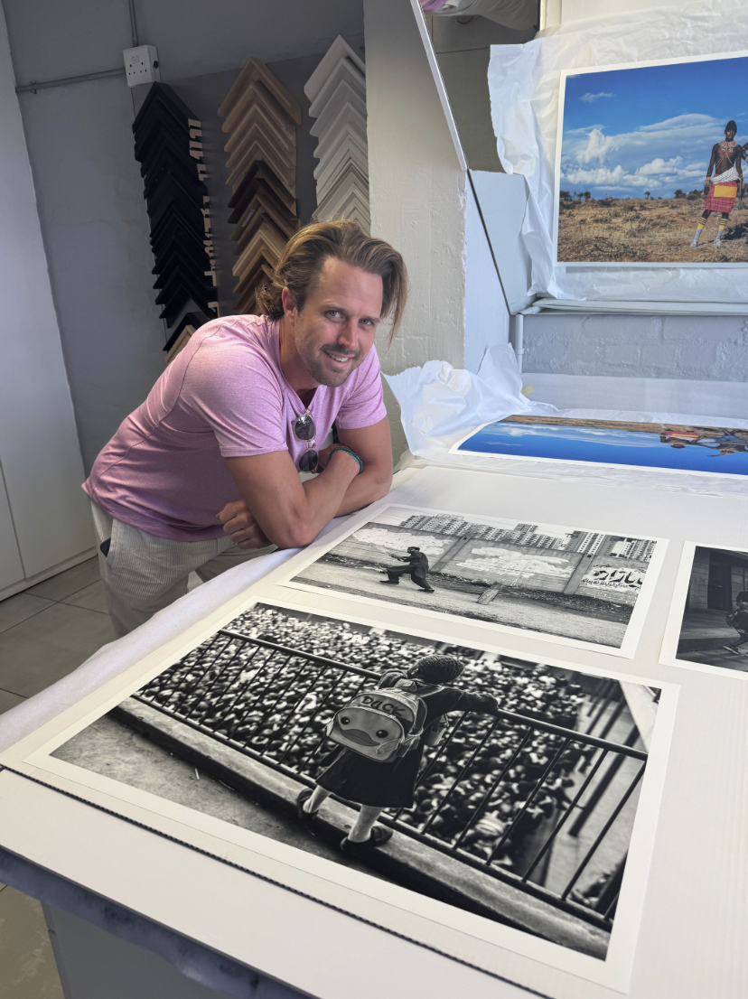 A man with light brown hair and a pink T-shirt leaning on a table displaying large photographs in a framing shop. The photos include a large black and white close-up of a child with a duck backpack looking over a railing, a man jumping in front of ci