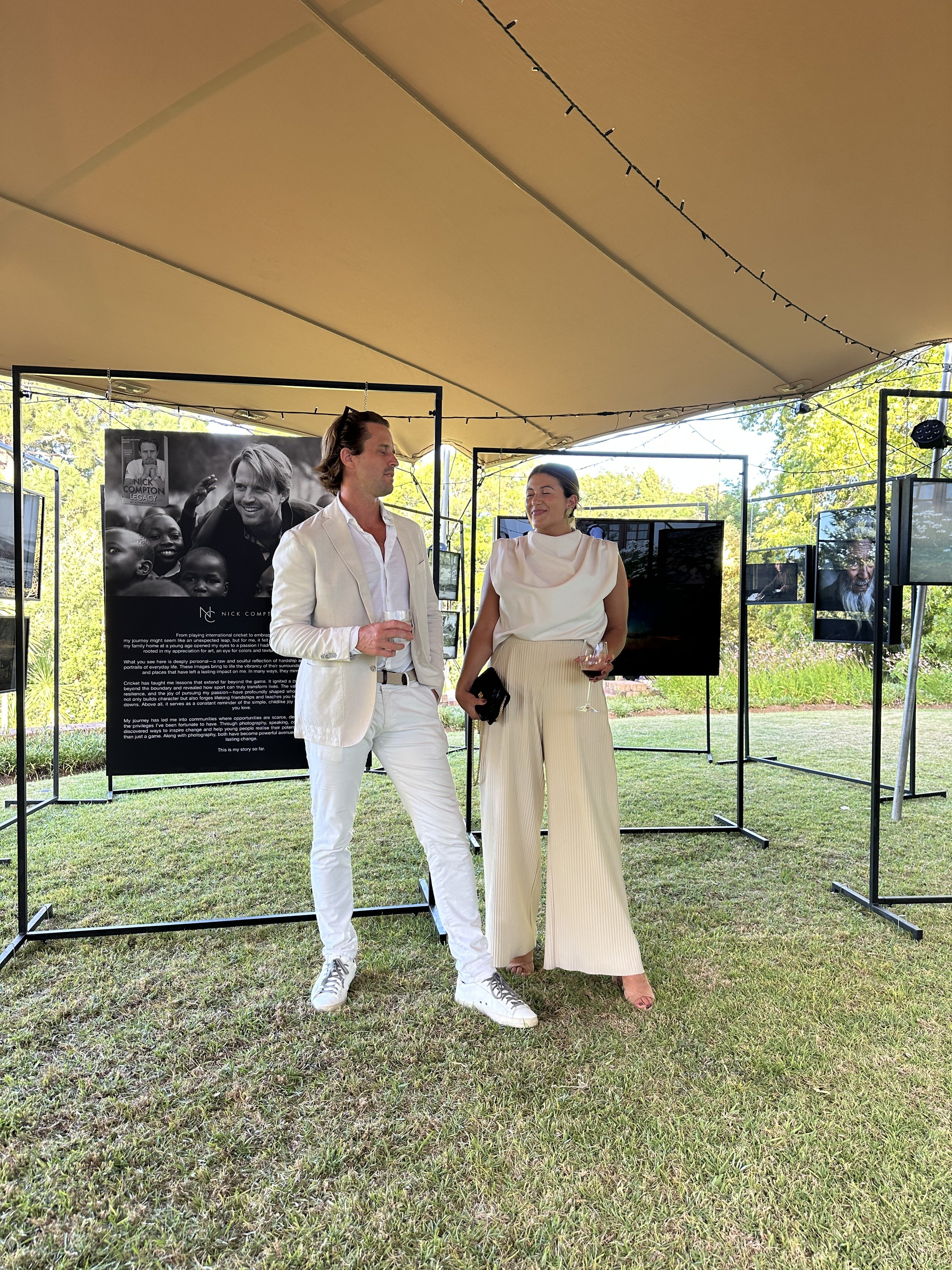 A man and woman are standing outdoors under a tent, dressed in white and cream-colored outfits, holding glasses of wine, and engaging in conversation at an art exhibit with black and white photographs displayed on black frames.