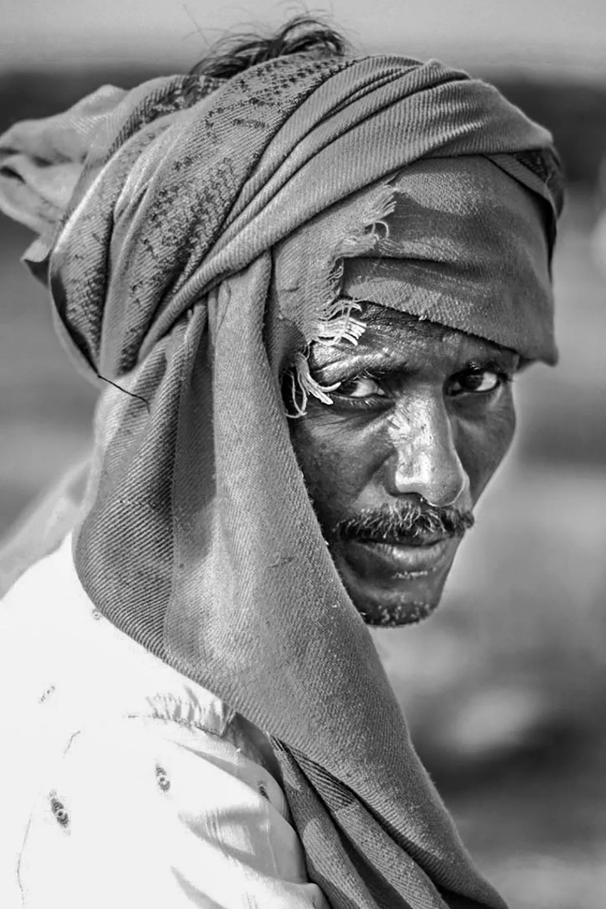 Black and white portrait of a man with a headscarf, looking directly at the camera with intense eyes.