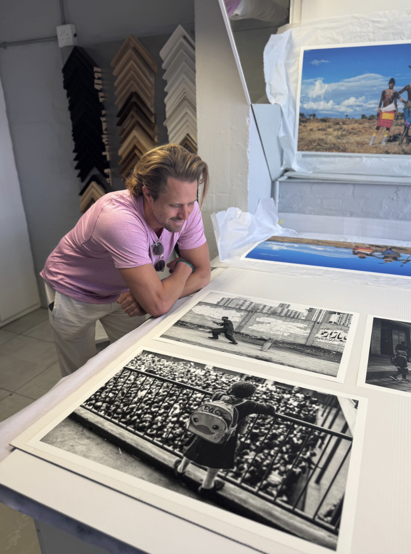 A man with shoulder-length hair wearing a pink t-shirt and sunglasses looks at photographs on a table in an art gallery or framing shop. There are framed photos on the table, including a black-and-white photo of a child in a backpack standing by a railing, and another of a person riding a bike on a city street. Framed art or photographs hang on the wall, including a large colorful landscape photograph of two people in a rural setting.
