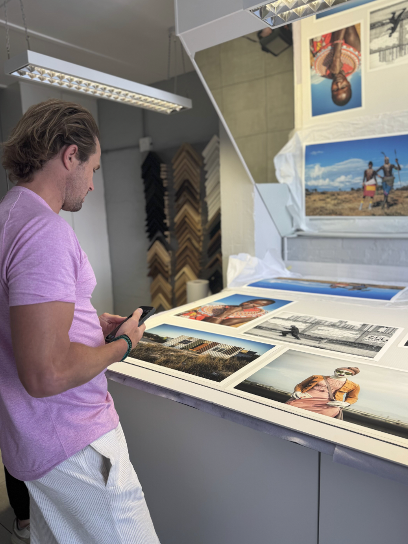 A man in a pink shirt and white striped pants looks at photographs laid out on a table in a gallery or framing shop.