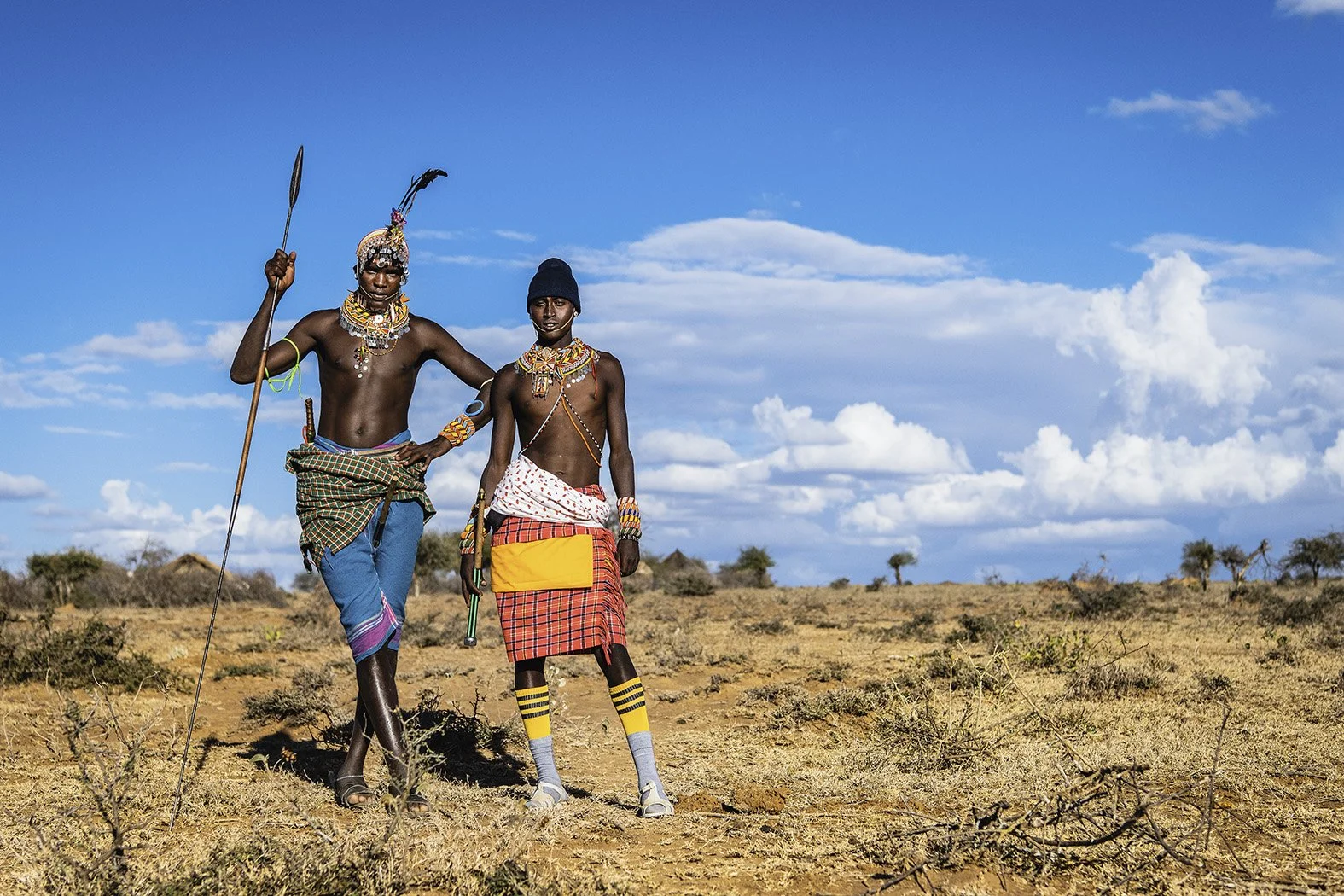 Two men dressed in traditional Maasai attire standing in a dry, open landscape with scattered bushes and trees under a partly cloudy sky.