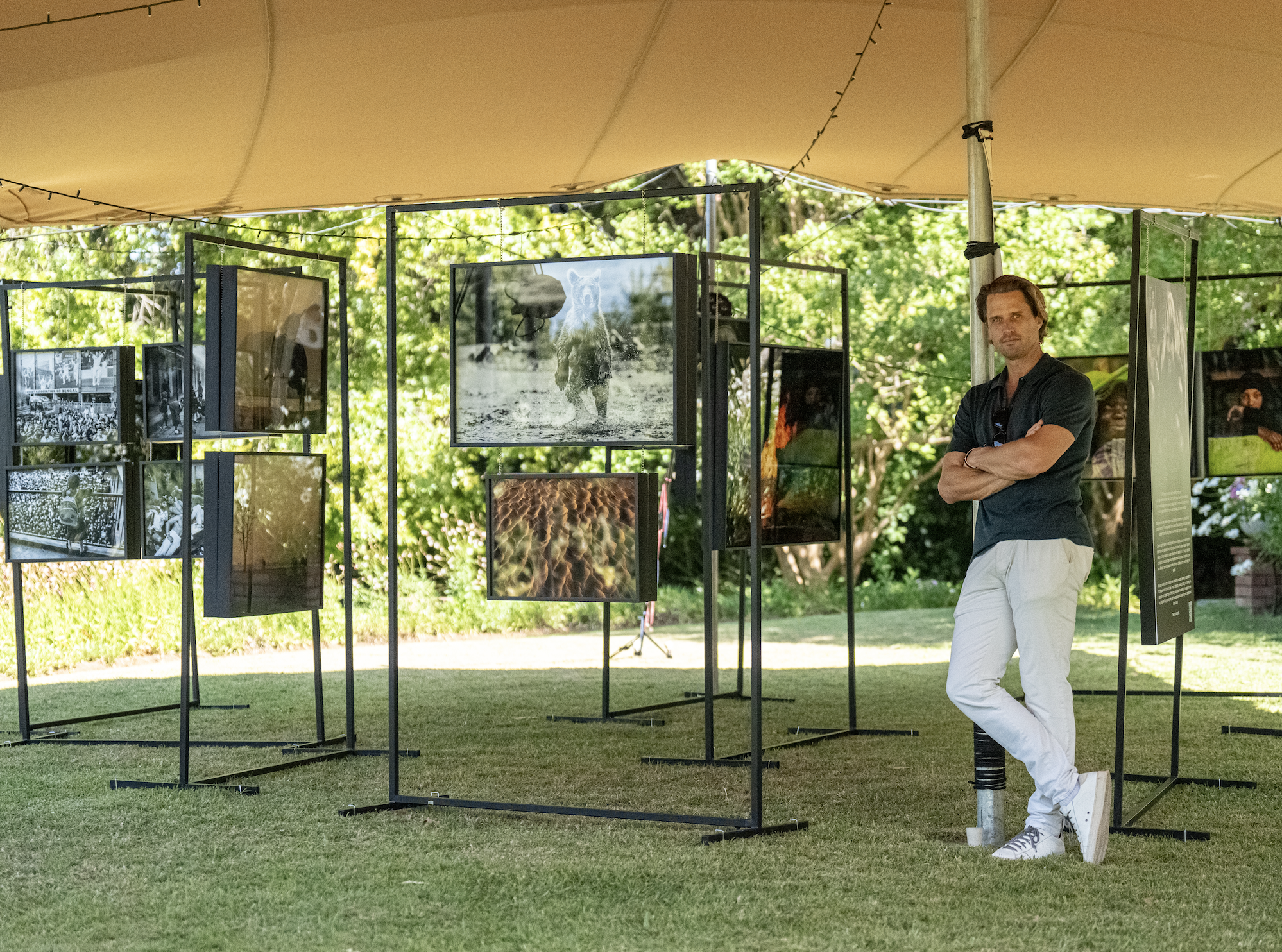 A man in white pants and a dark shirt stands with arms crossed in front of an outdoor photo exhibition under a tent, with panels displaying various photographs of animals and nature.