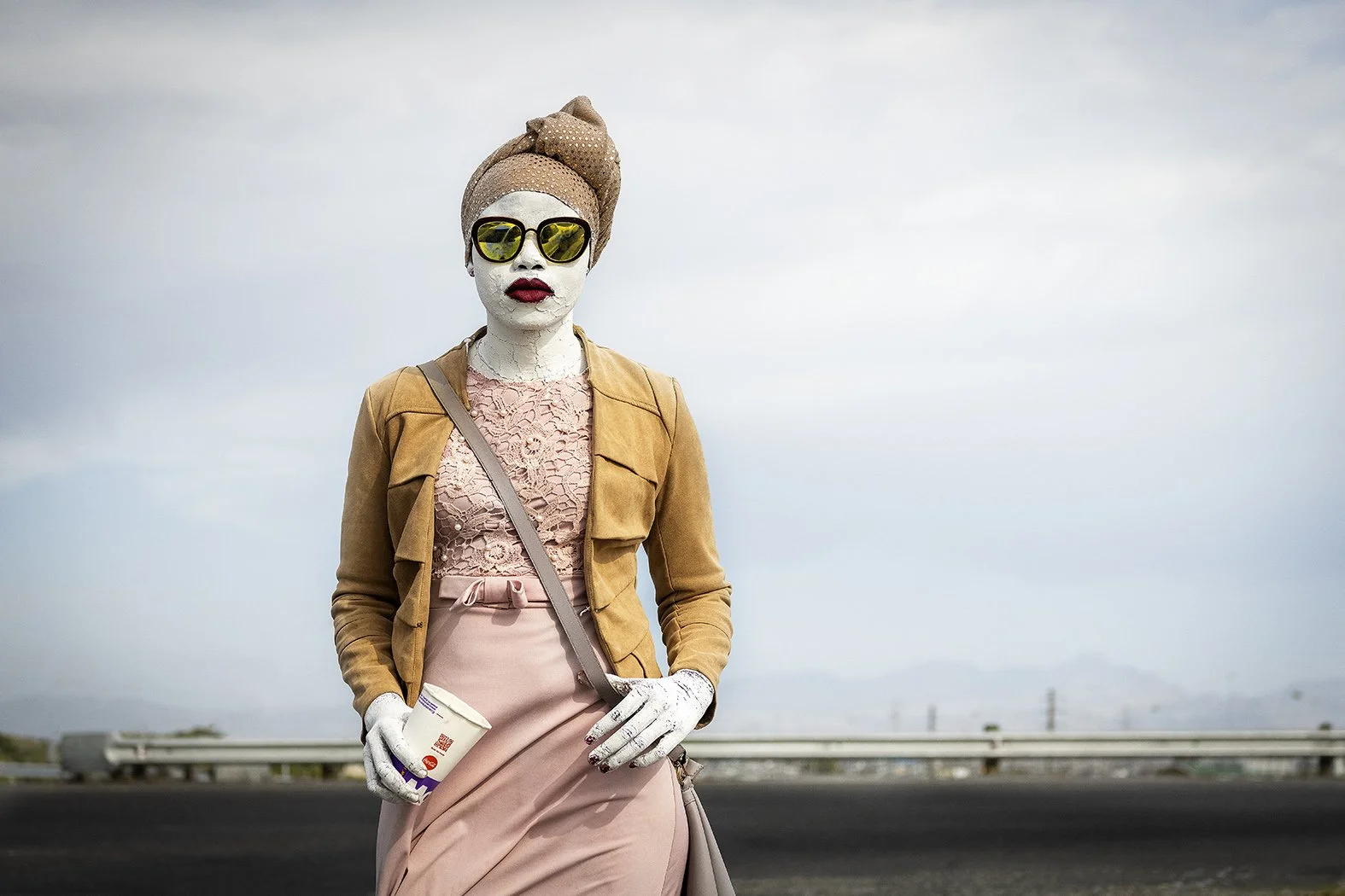 A woman with painted white face makeup, wearing large sunglasses, a pink lace top, a tan jacket, and a pink skirt, standing outdoors on a cloudy day, holding a coffee cup.