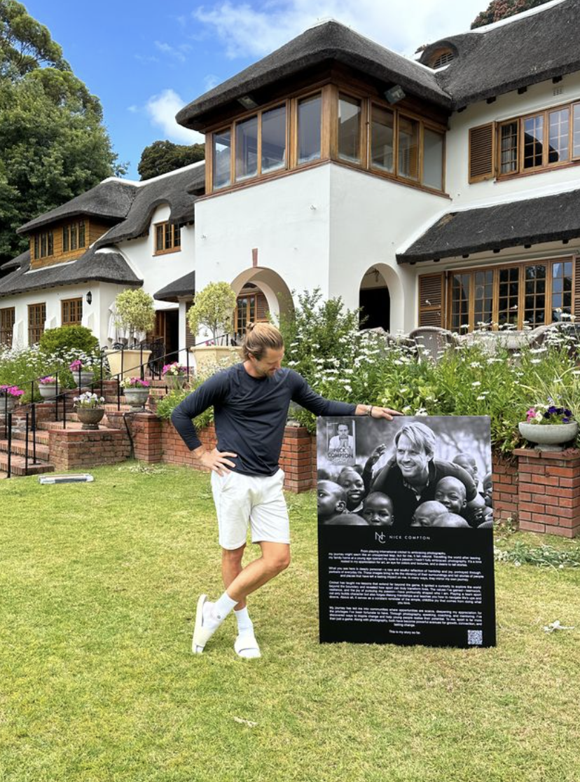A person standing on a lawn, touching a black and white informational sign with a photo of a man and children. The background shows a large white house with wooden-framed windows, a thatched roof, and a garden with flowers and trees.