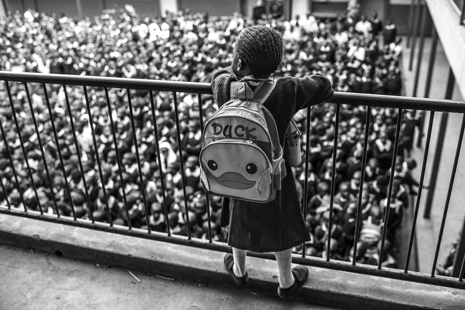 A young girl wearing a backpack with a duck face design, standing at a balcony and looking out over a large crowd of students sitting in an auditorium.