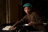 Young boy in brown traditional clothing and a blue hairnet wiping the surface of a dark kitchen counter with a cloth, holding a metal bowl in his other hand.