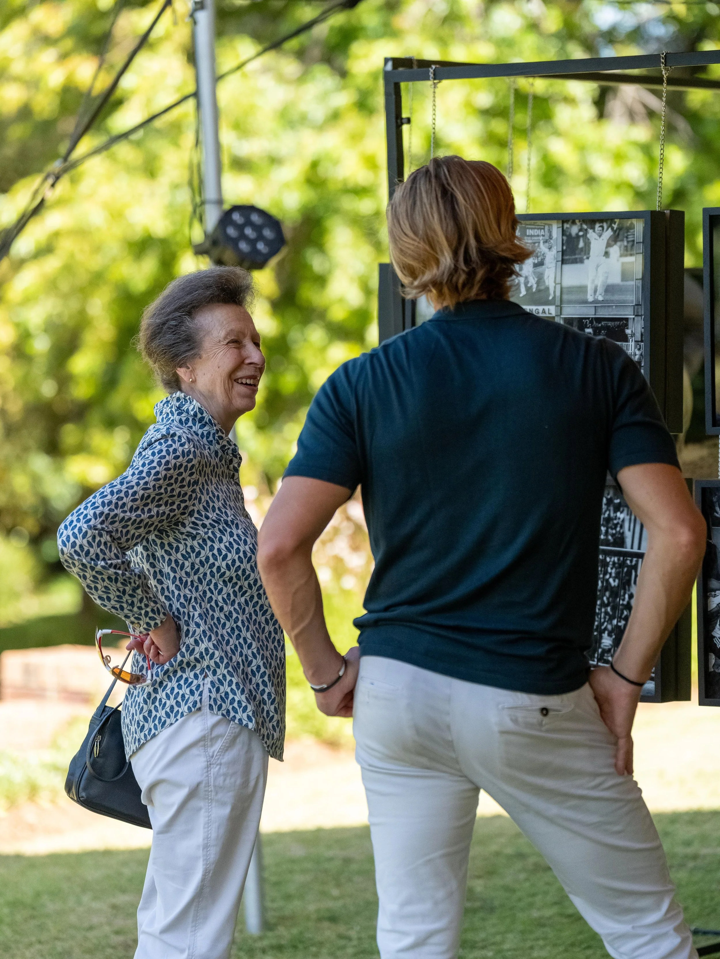 An elderly woman and a younger woman are standing outdoors, engaging in conversation and smiling. The elderly woman is wearing a patterned blouse and white pants. The younger woman is dressed in a navy shirt and white pants, with her back to the came