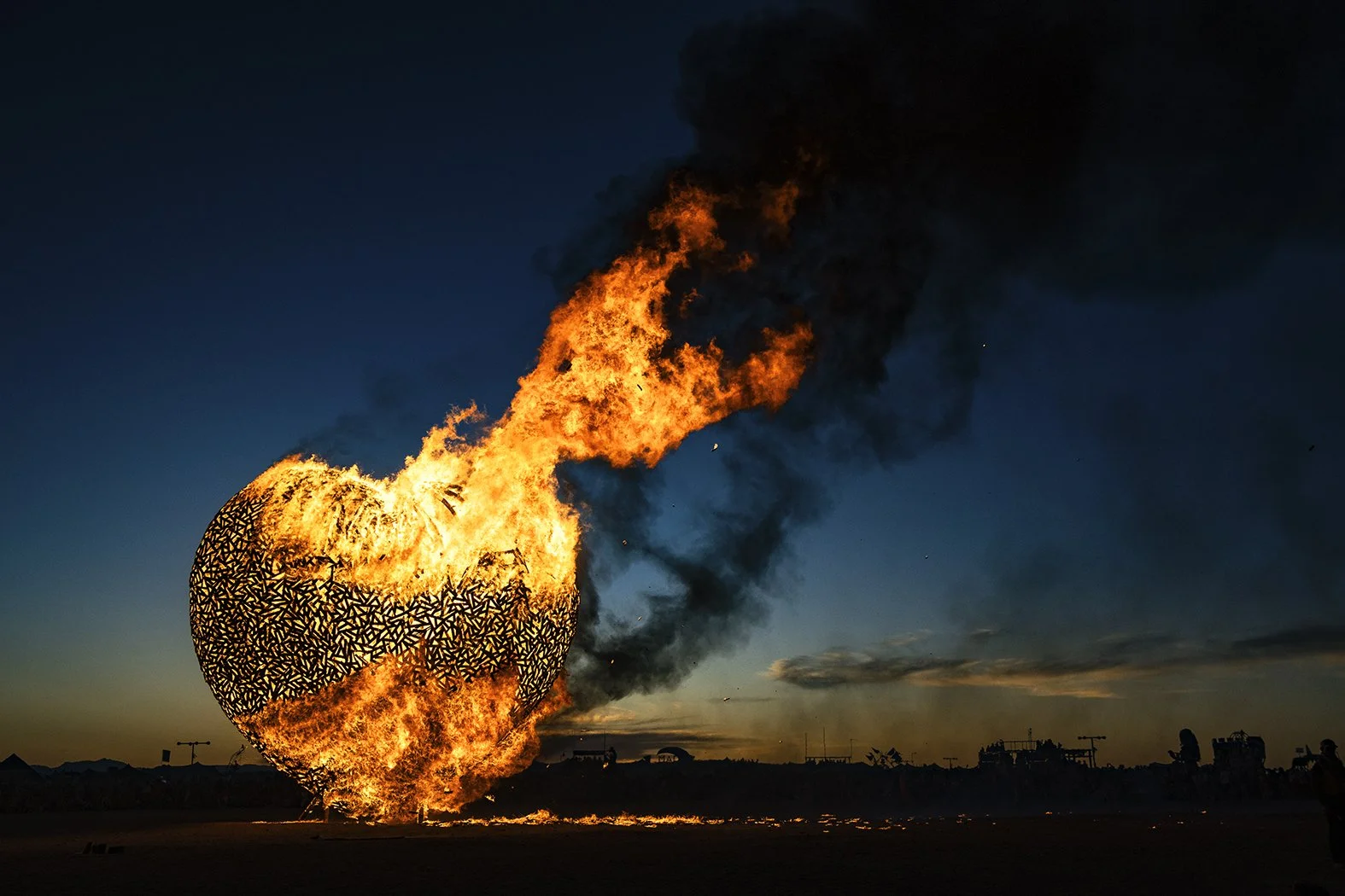 A burning heat balloon in the shape of a heart with black geometric patterns on its surface, erupting with flames and black smoke against a darkening sky.