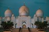 White mosque with multiple large domes, surrounded by palm trees, staircase leading up to the entrance, and lit at dusk.