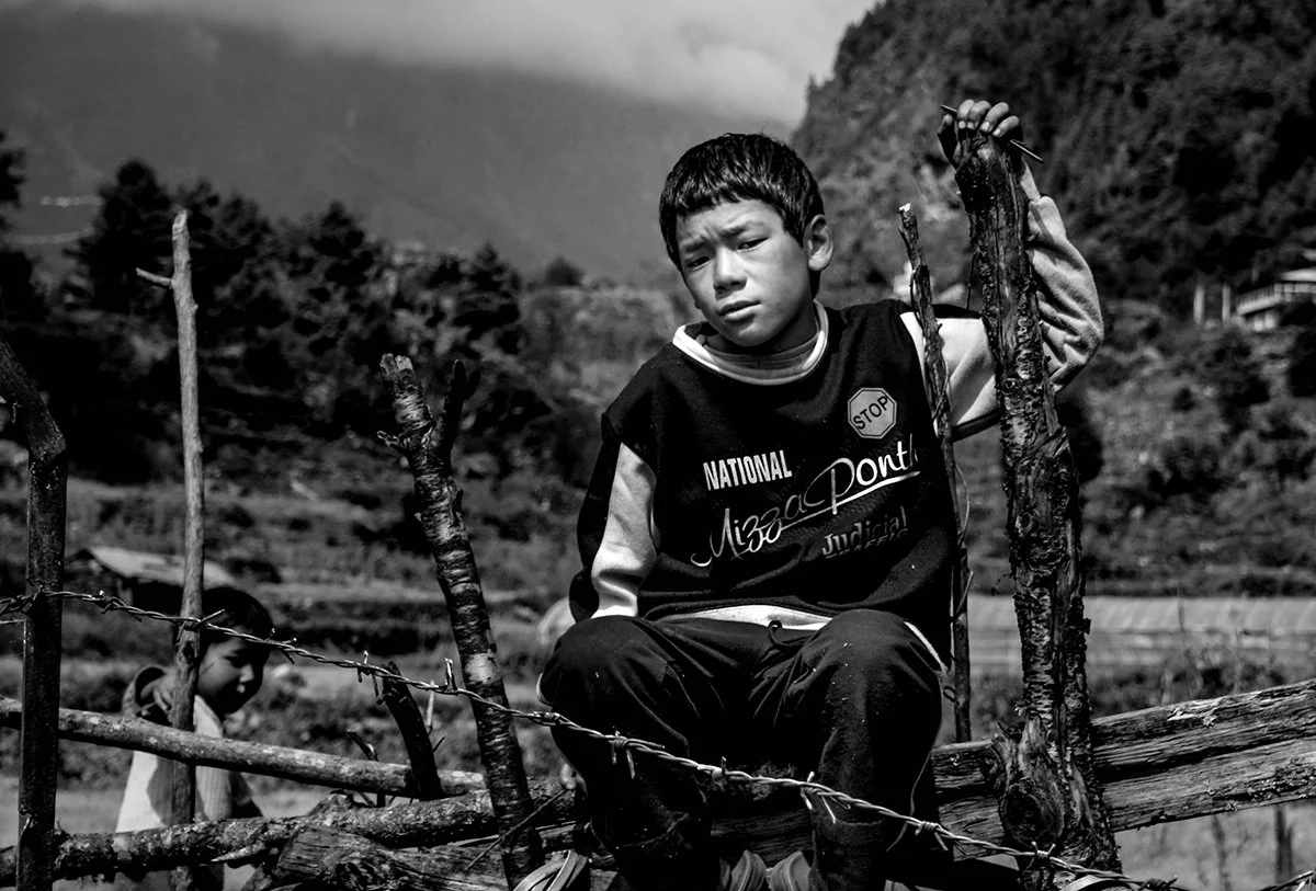 A young boy sitting on a wooden structure outside, holding two sticks, with mountains and trees in the background.