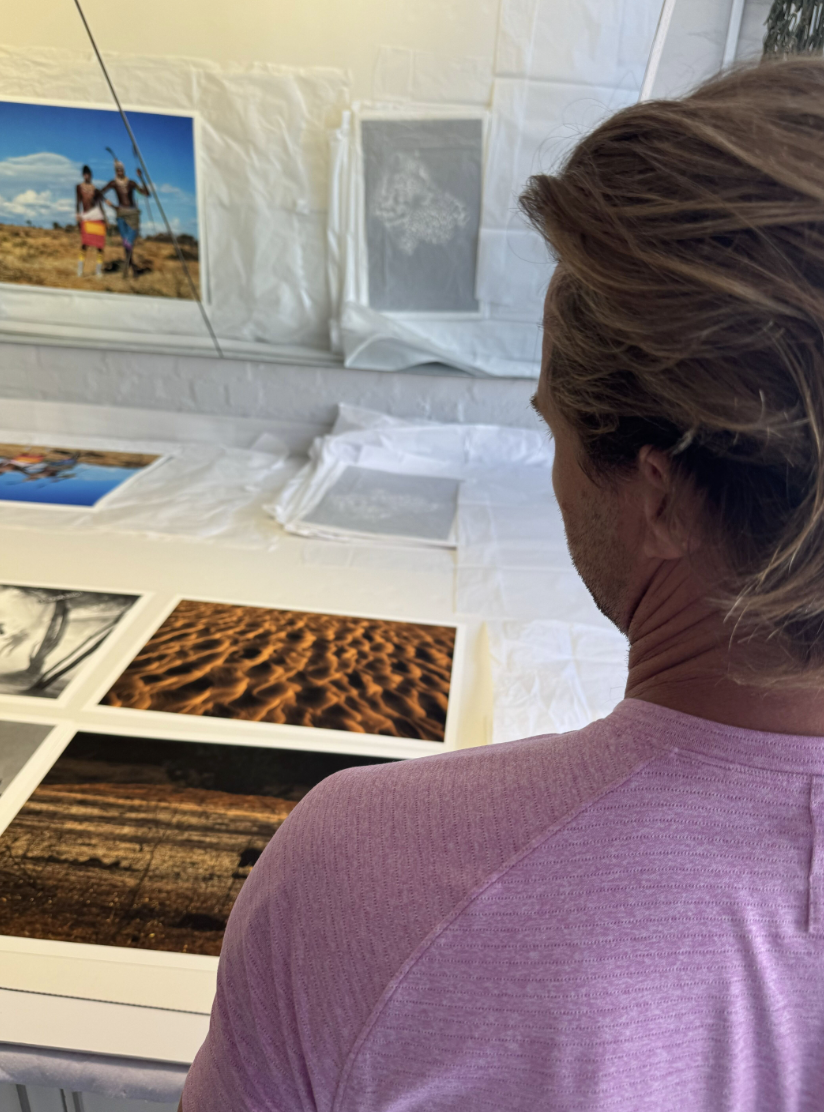 Person with long brown hair and wearing a pink shirt looking at printed photographs on a table in a white-walled room.