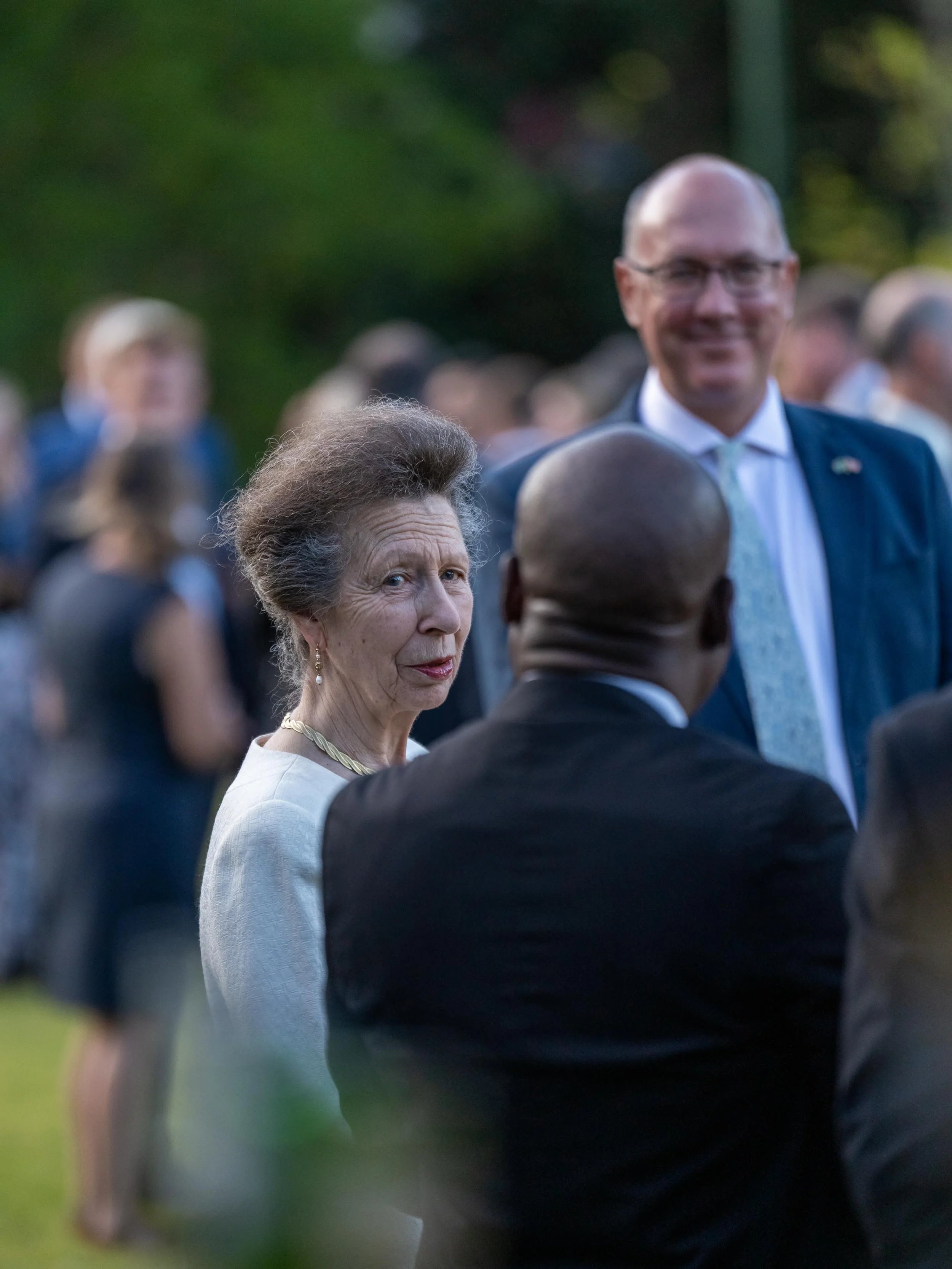 A woman with gray hair and a white outfit standing outdoors at a social event, engaging in conversation with a man with a bald head and dark suit, while a smiling man in a suit and tie stands in the background.