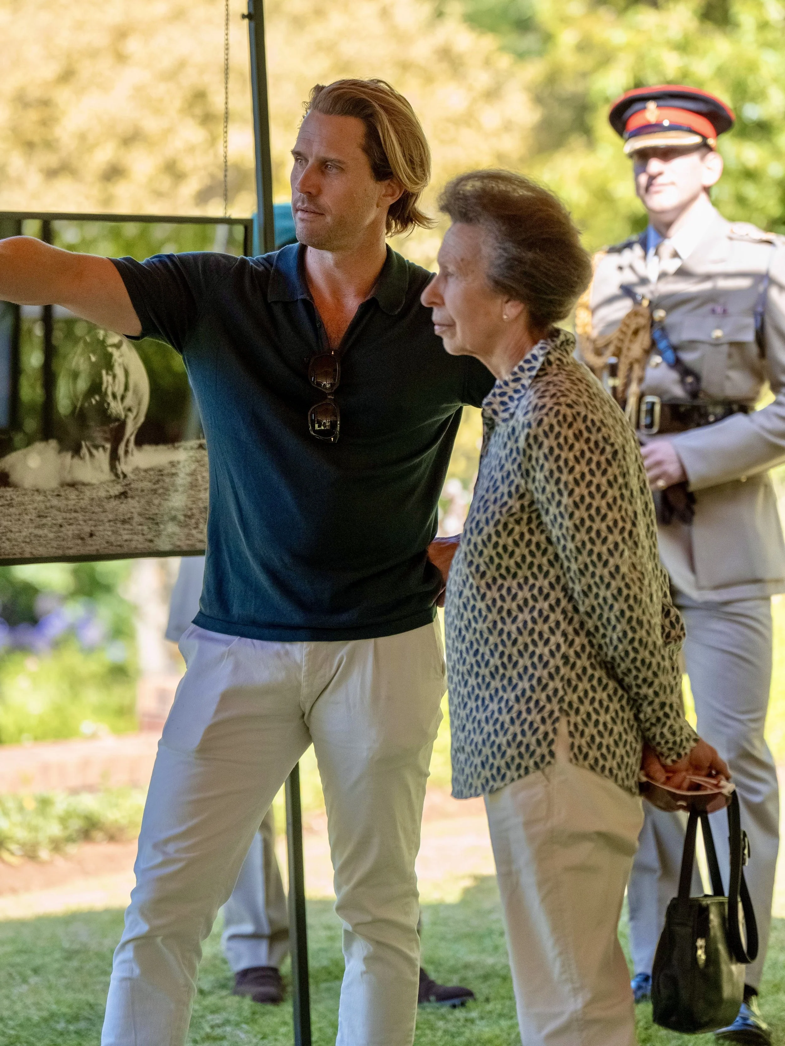 A young man in a black polo shirt and cream pants pointing at a picture displayed on an easel, while an elderly woman in a patterned blouse and cream pants looks on, holding a small black bag, in an outdoor garden setting with trees and greenery. A m