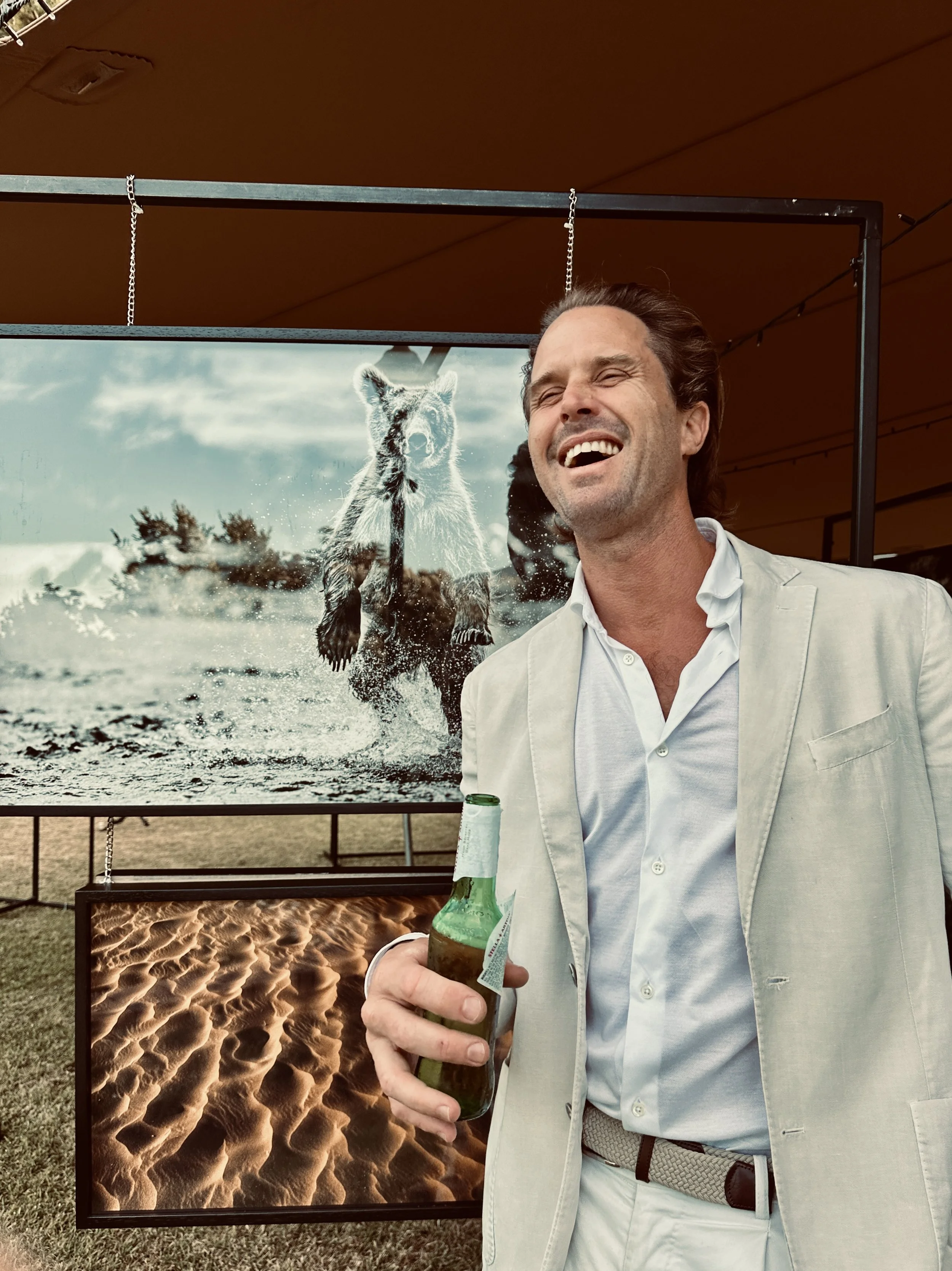 A man dressed in a light-colored suit holding a beer, smiling, standing outside in front of framed photographs including a racing wild animal and sandy terrain.