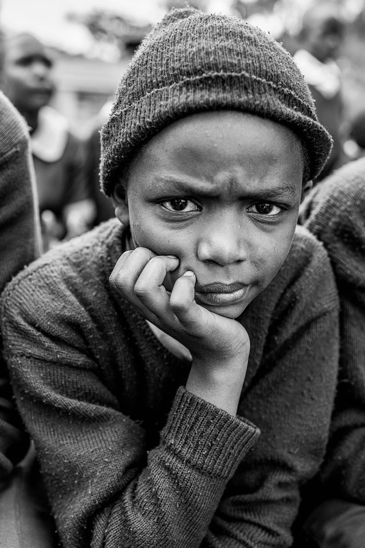 A black and white photo of a young boy wearing a beanie and sweater, resting his chin on his hand, looking directly at the camera with a serious expression.