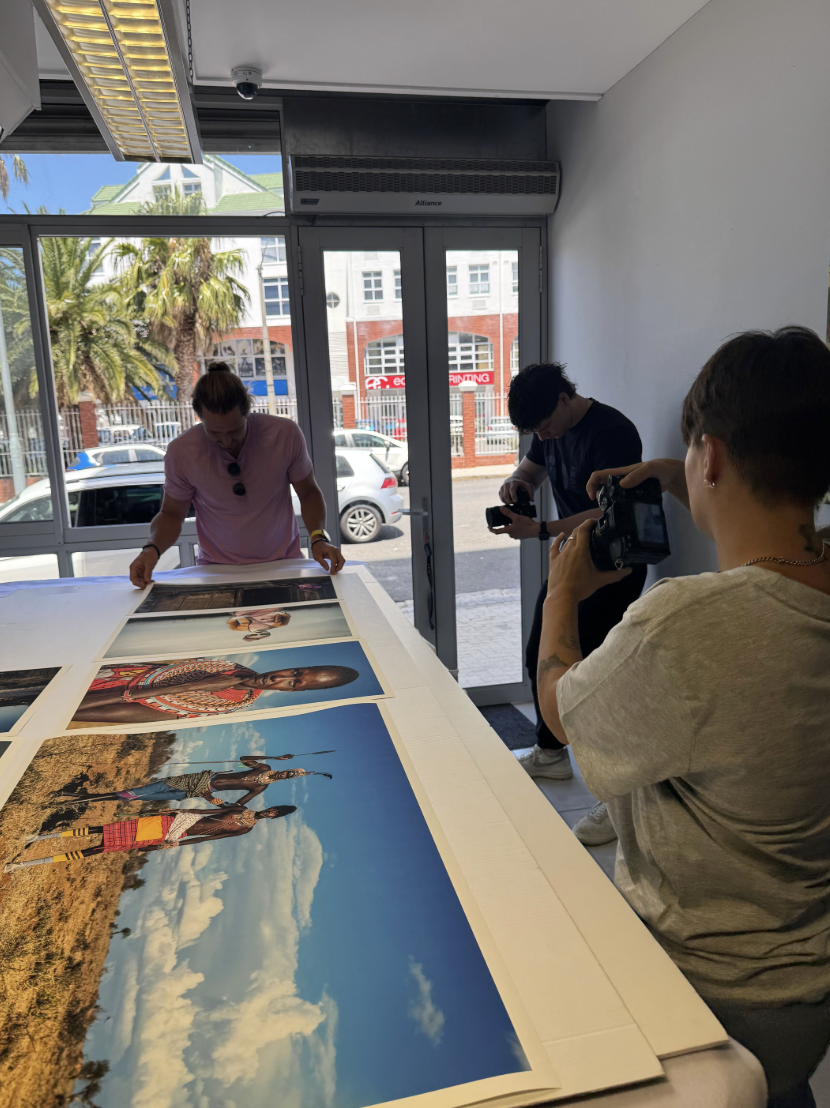 People looking at large printed photographs displayed on a table inside a room with glass doors and windows showing parked cars and trees outside.