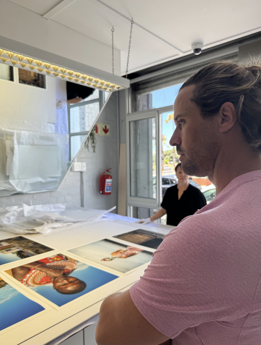 A man with long hair wearing a pink shirt is standing indoors, looking at photographs laid out on a table. Behind him, a woman in a black top stands near a window, and there is a fire extinguisher on the wall.