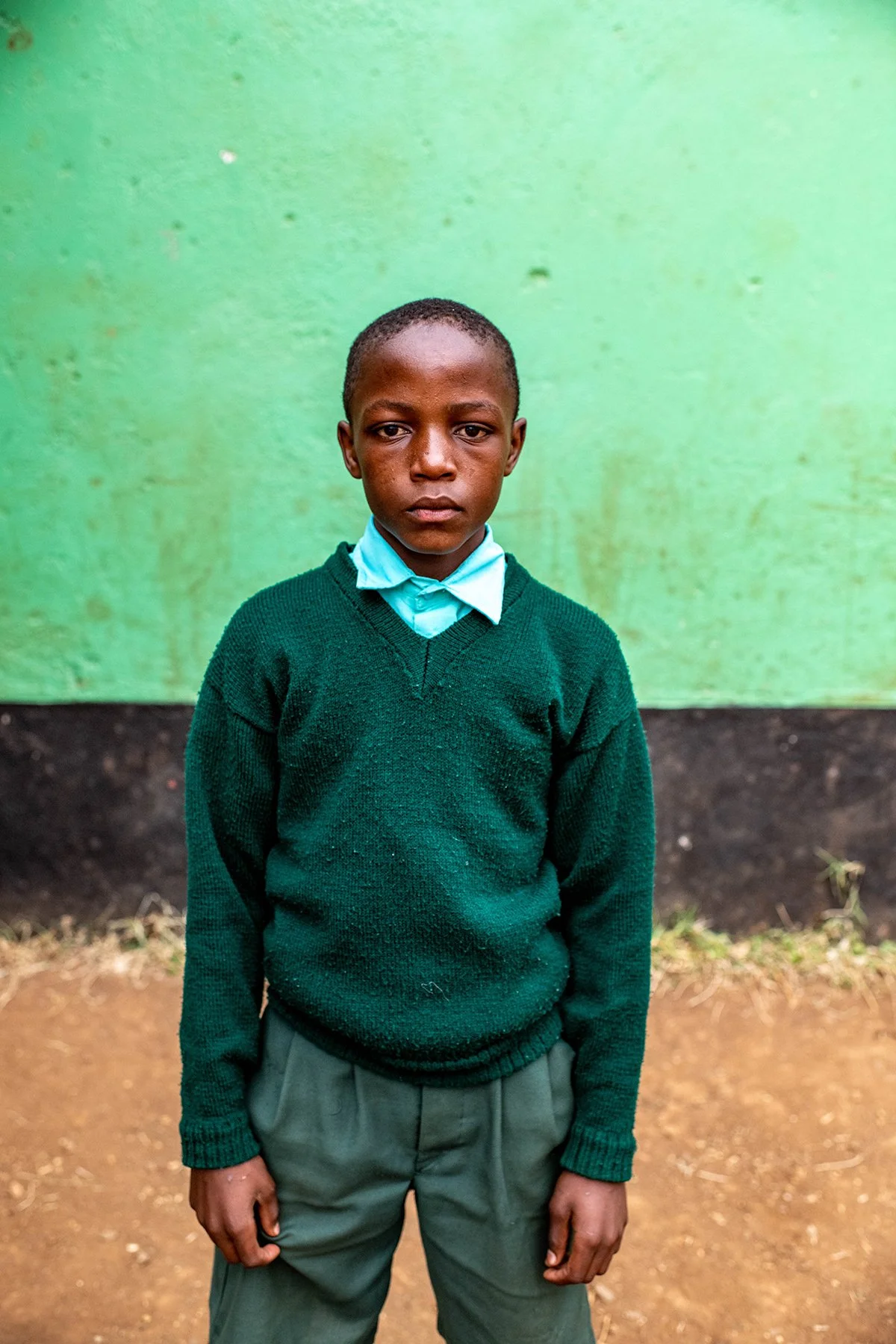 A young boy in school uniform standing in front of a green wall with a somber expression.