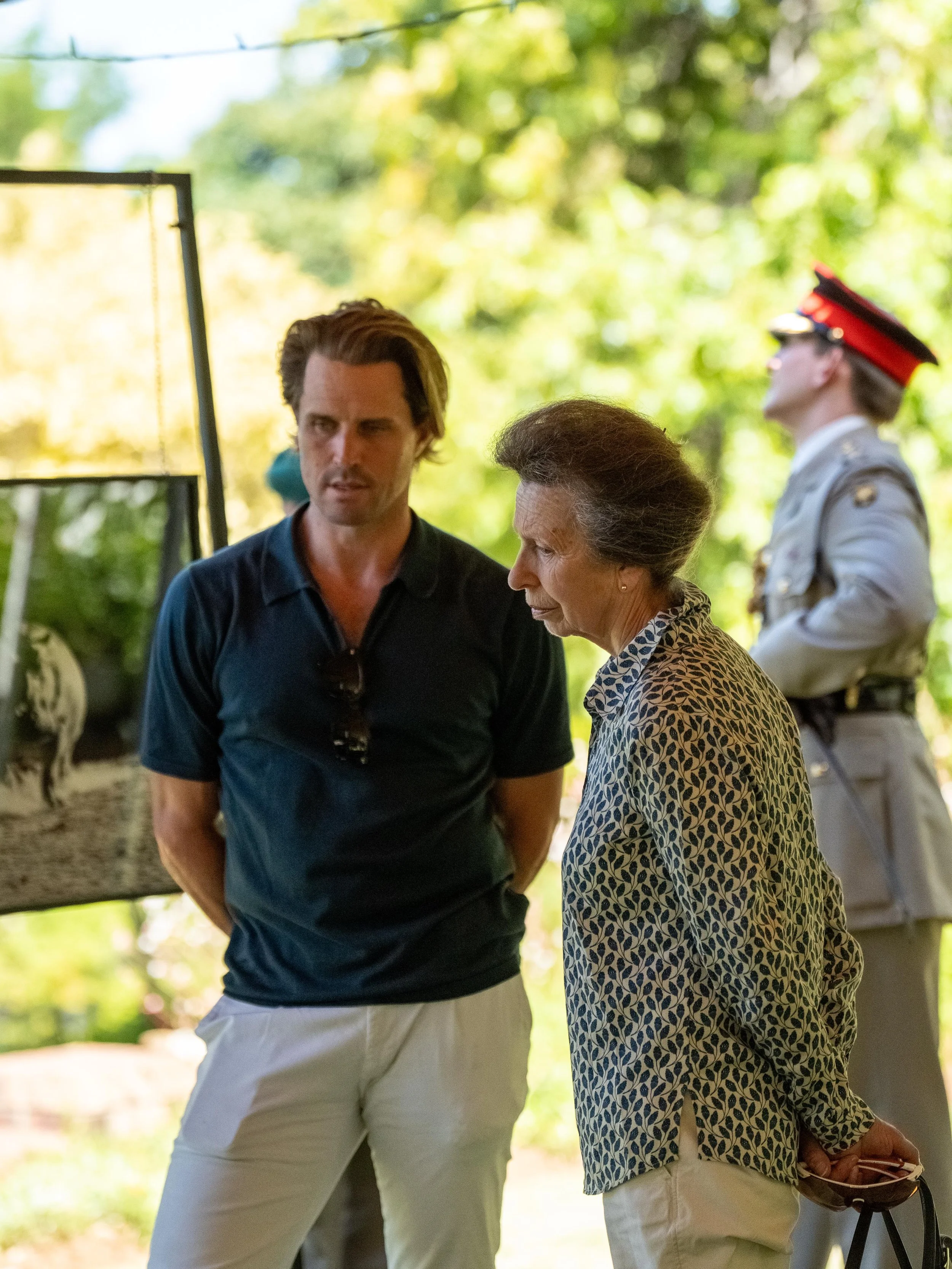 A man and an elderly woman looking at something outdoors, with a person in a military uniform standing in the background.