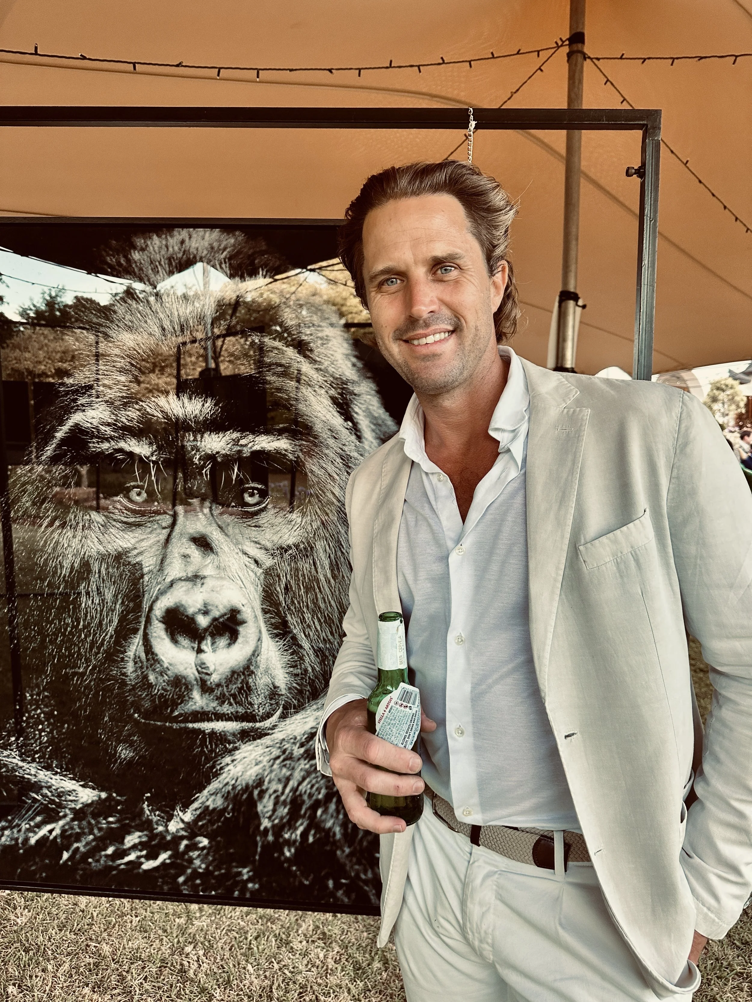 A man in a white suit holding a beer stands next to a large black and white photograph of a gorilla with a waterfall in the background.
