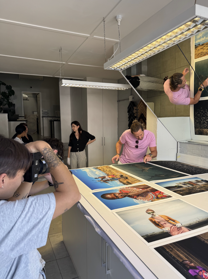 People examining large printed photographs on a table in an art gallery or studio, with a mirror on the wall reflecting the scene.