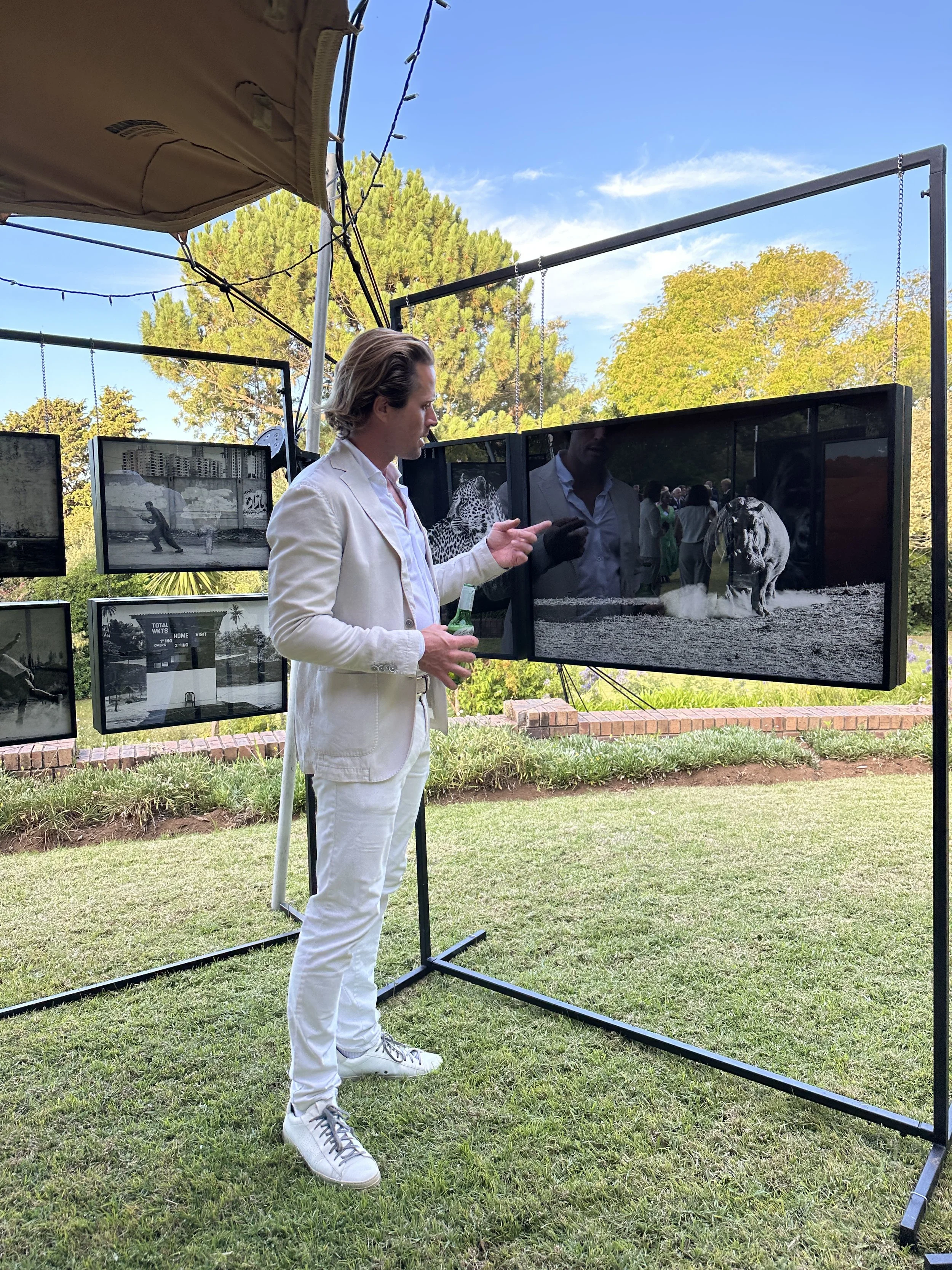 A man in a white suit and sneakers stands outside looking at a display of black-and-white photographs of animals on hanging panels, with trees and blue sky in the background.