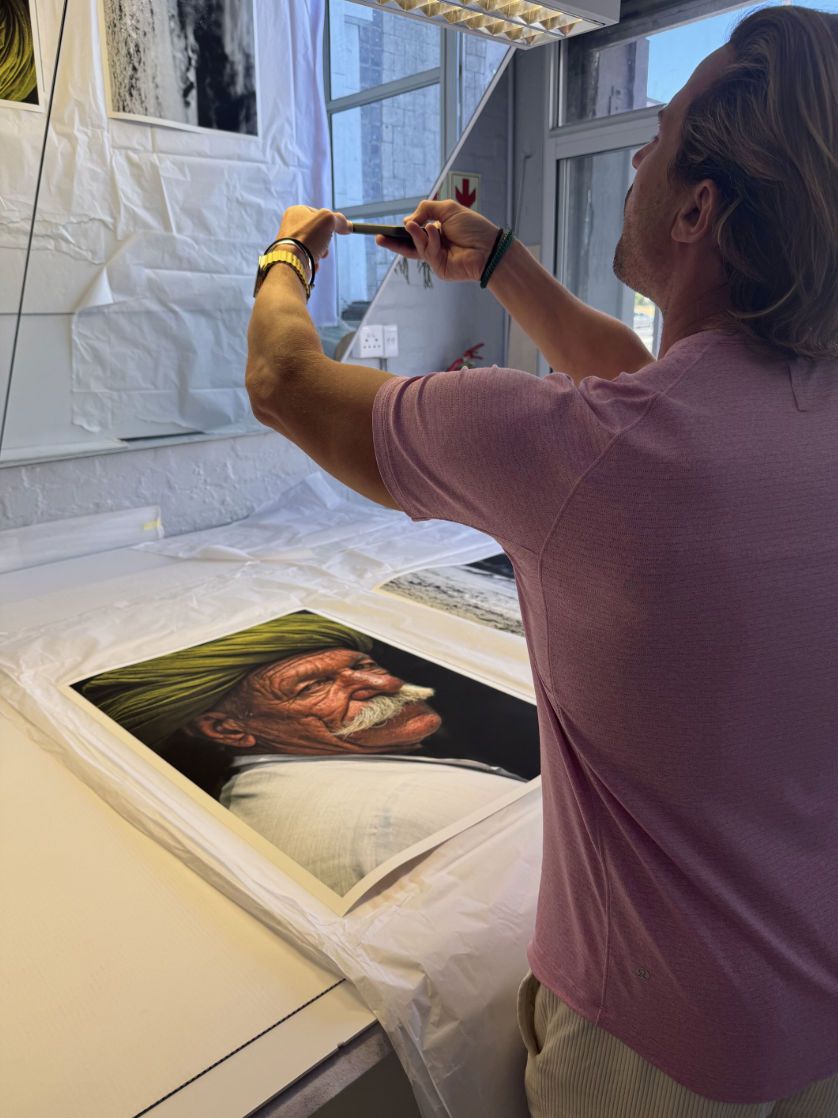 A man with shoulder-length hair taking a photograph of a large portrait of an elderly man with a white mustache wearing a green turban and white shirt, lying on a table in an art studio with large windows.