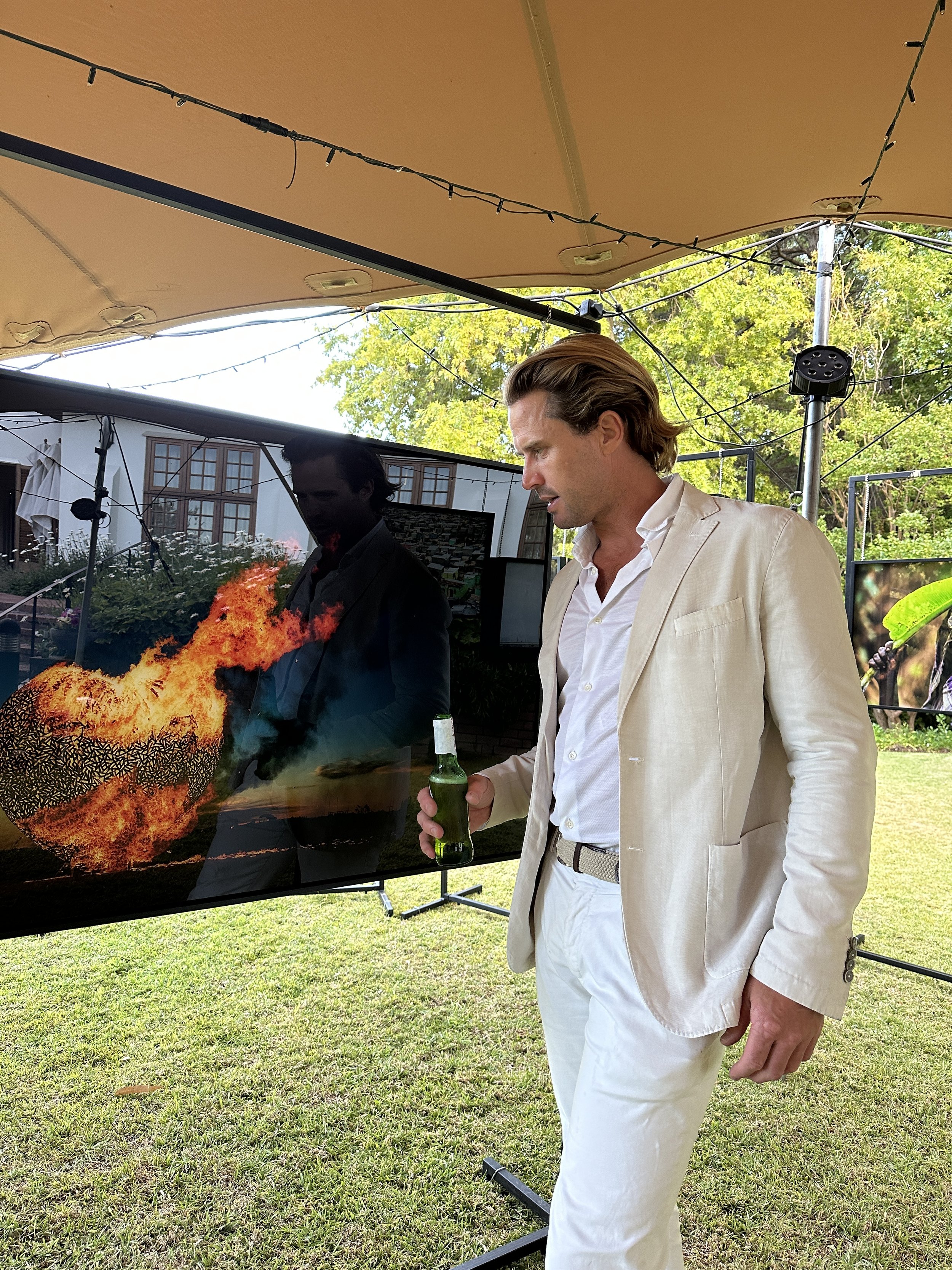 A man in a cream-colored suit holding a beer bottle observes a screen showing a fire and a fireball. The setting is outdoors under a tan canopy with string lights. There are trees and a house in the background.