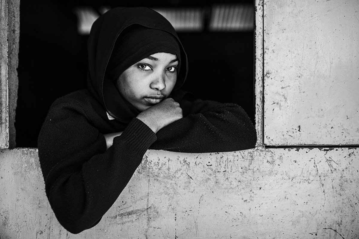 Black and white photo of a young woman with a hijab resting her chin on her crossed arms, looking thoughtfully out of a concrete window opening.