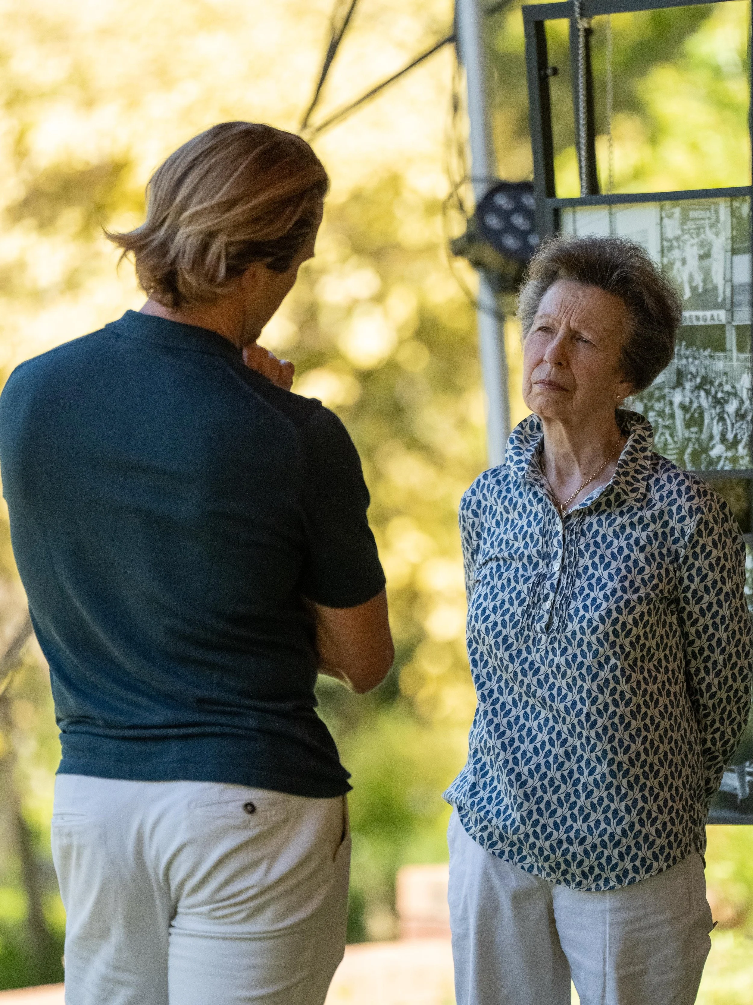 Two women are having a conversation outdoors with trees and a display board in the background.