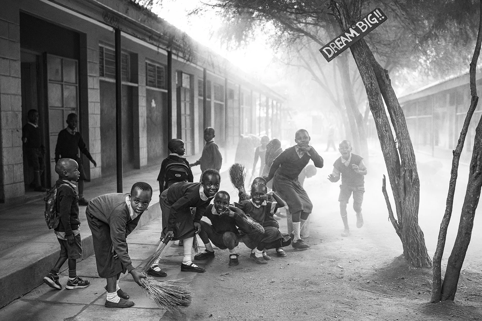 School children in uniforms playing and running outside on a dirt path near a school building, with a tree and a sign that reads 'DREAM BIG!!' in the foreground, all in black-and-white.