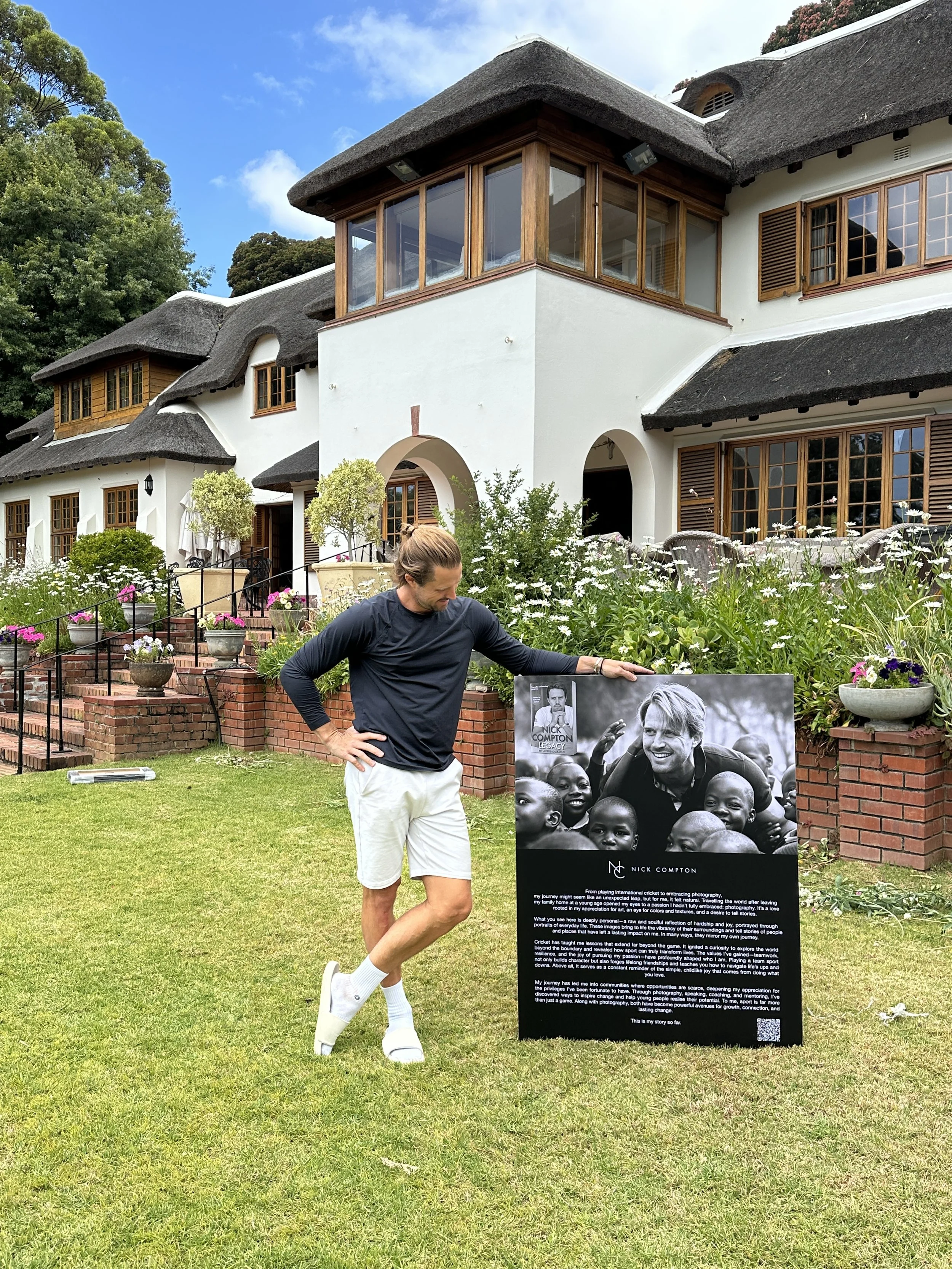 A man stands on a lawn in front of a house with white walls, wooden windows, and a thatched roof, looking at a black and white poster of a person surrounded by children.