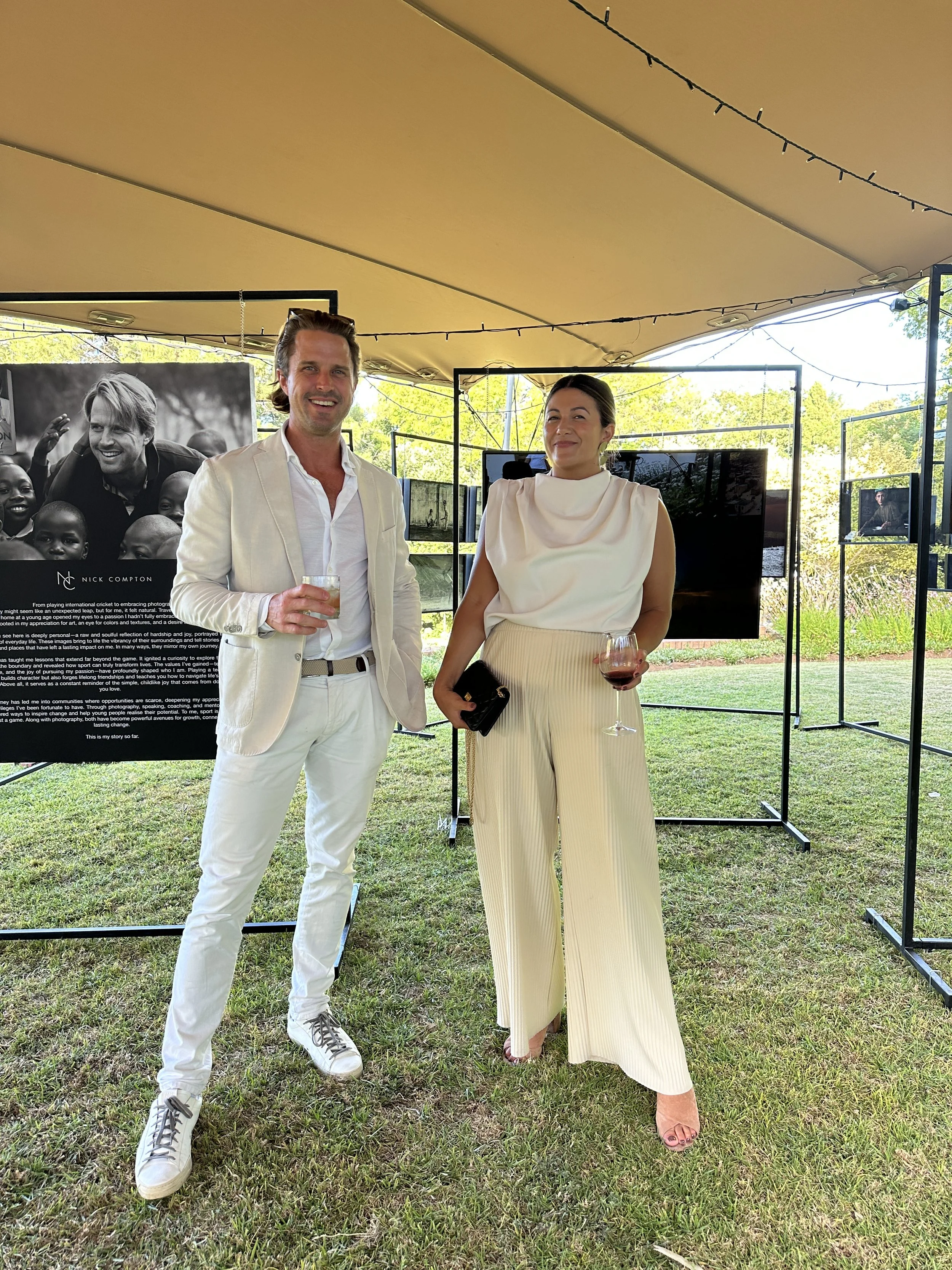 A man and woman dressed in white suits are standing outdoors at an art or photography exhibition, holding glasses of wine, with display panels and greenery in the background.