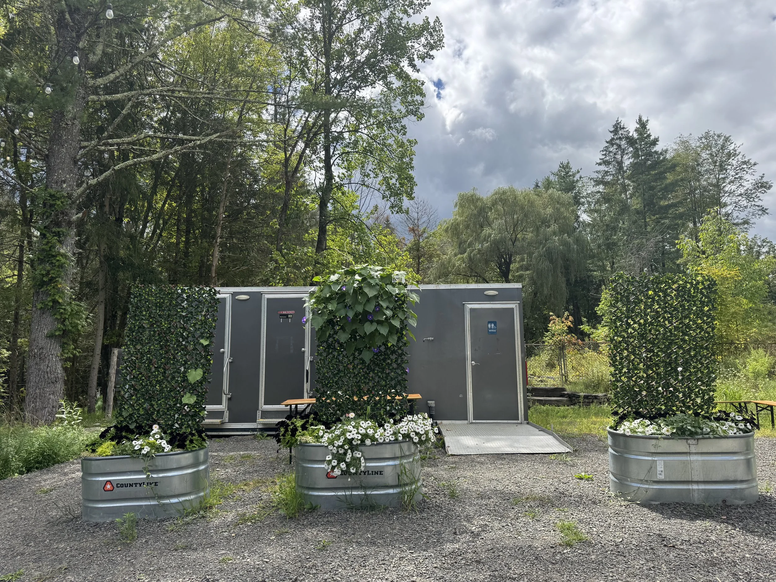 Portable restroom trailer with three doors, each with steps and railings, and potted flowers in front.
