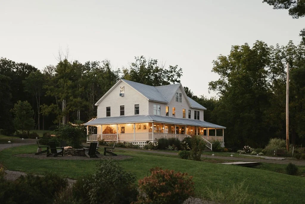 A large white farmhouse with a metal roof and wraparound porch, surrounded by greenery and trees.