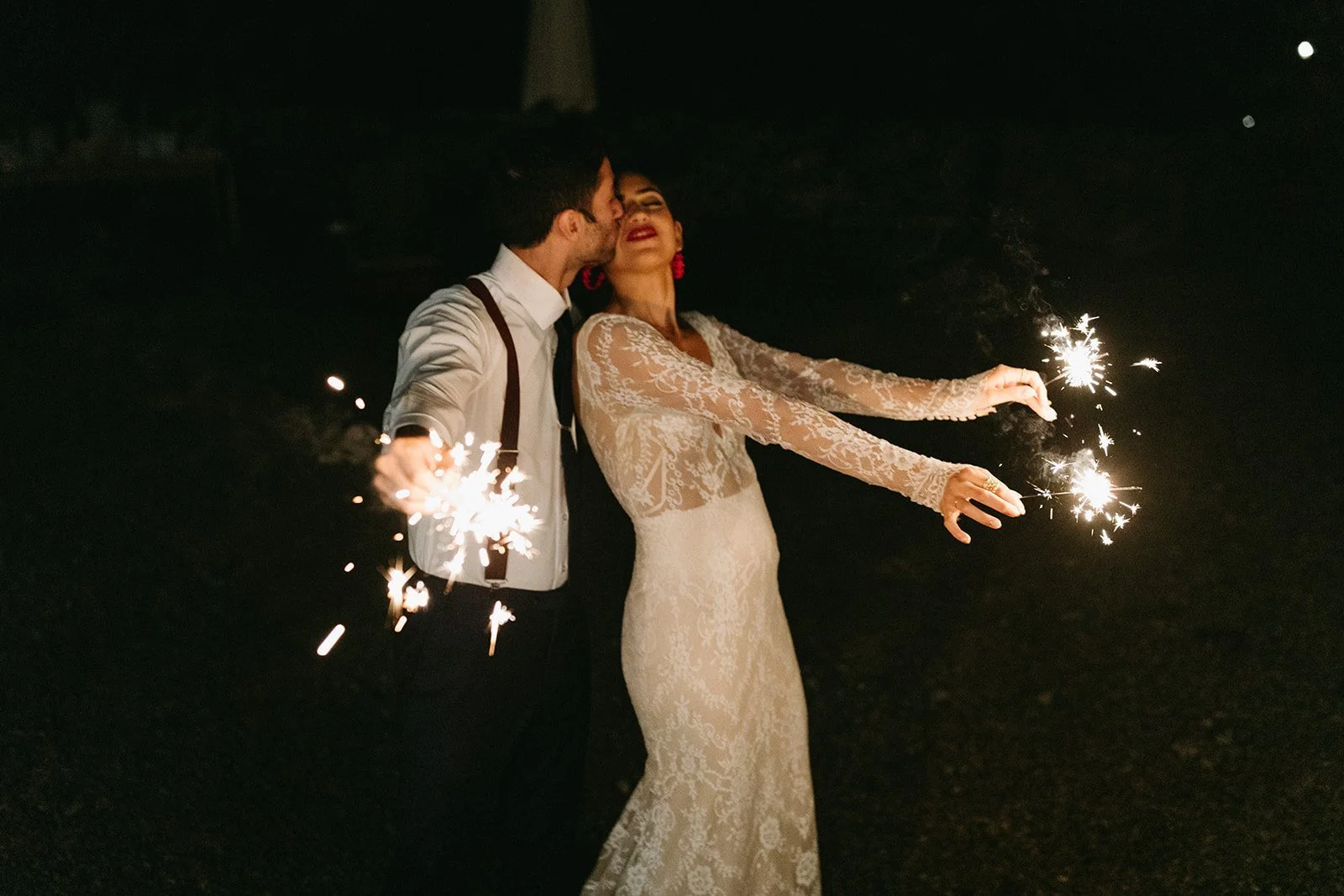 Bride and groom celebrating with sparklers at night, bride in a lace wedding dress and groom in a white shirt with suspenders.