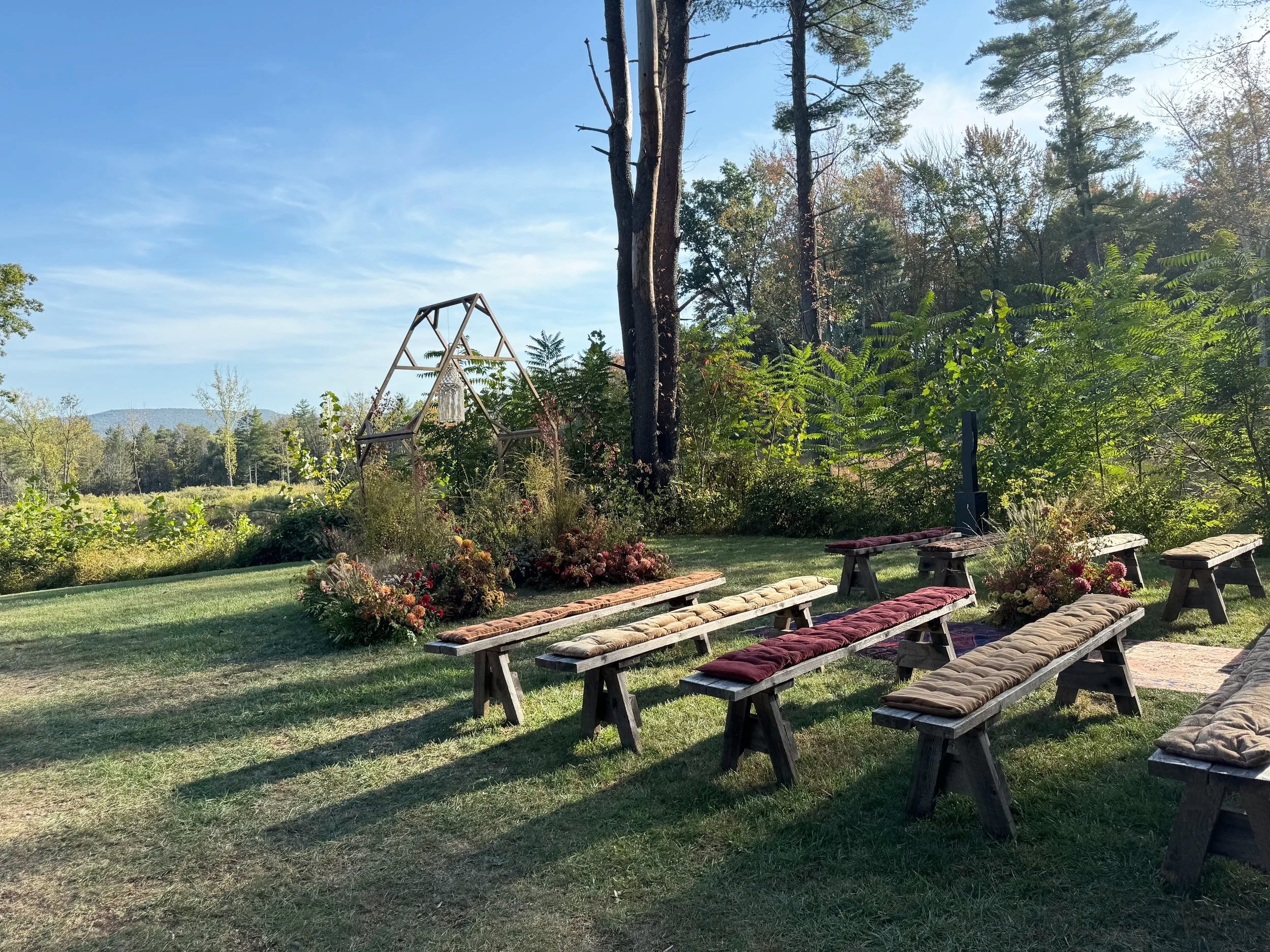 Outdoor wedding ceremony setup in a wooded area with wooden benches and pampas grass decorations.