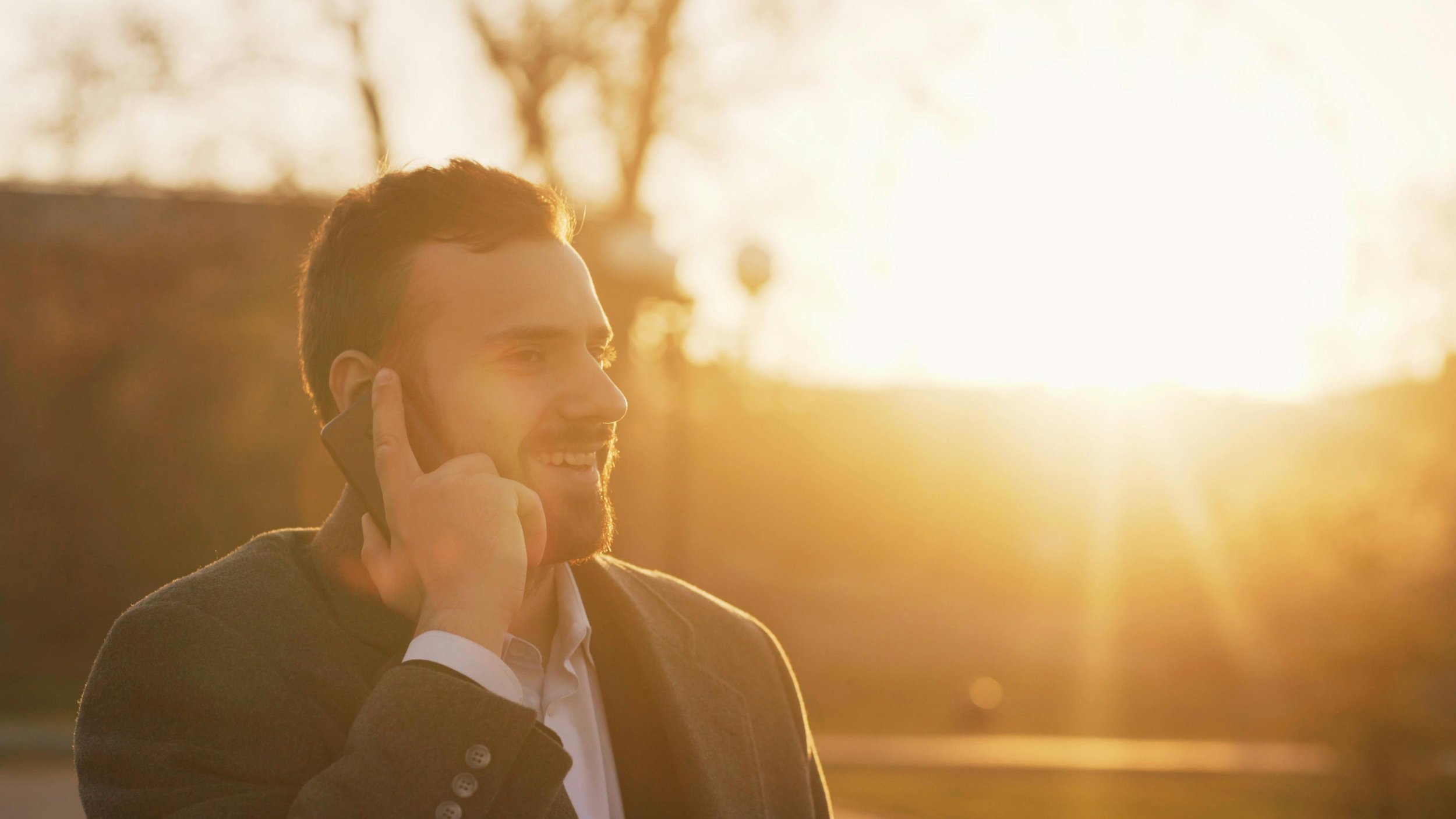 A man with short hair and a beard, wearing a dark blazer over a white shirt, is smiling and talking on a cellphone outdoors during sunset.