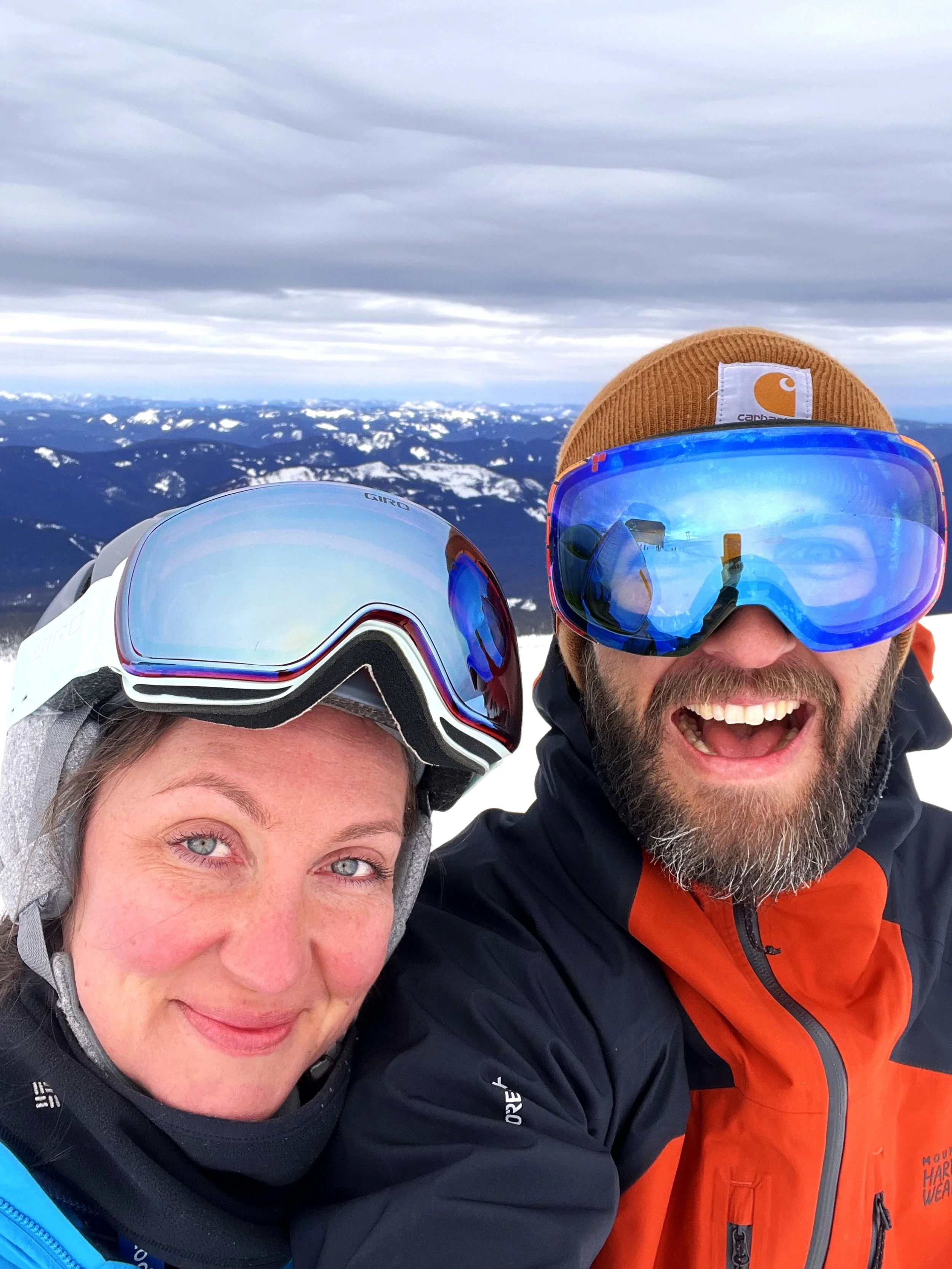 Two people in winter clothing and ski goggles are smiling for a selfie at high altitude, with snow-covered mountains and cloudy sky in the background.