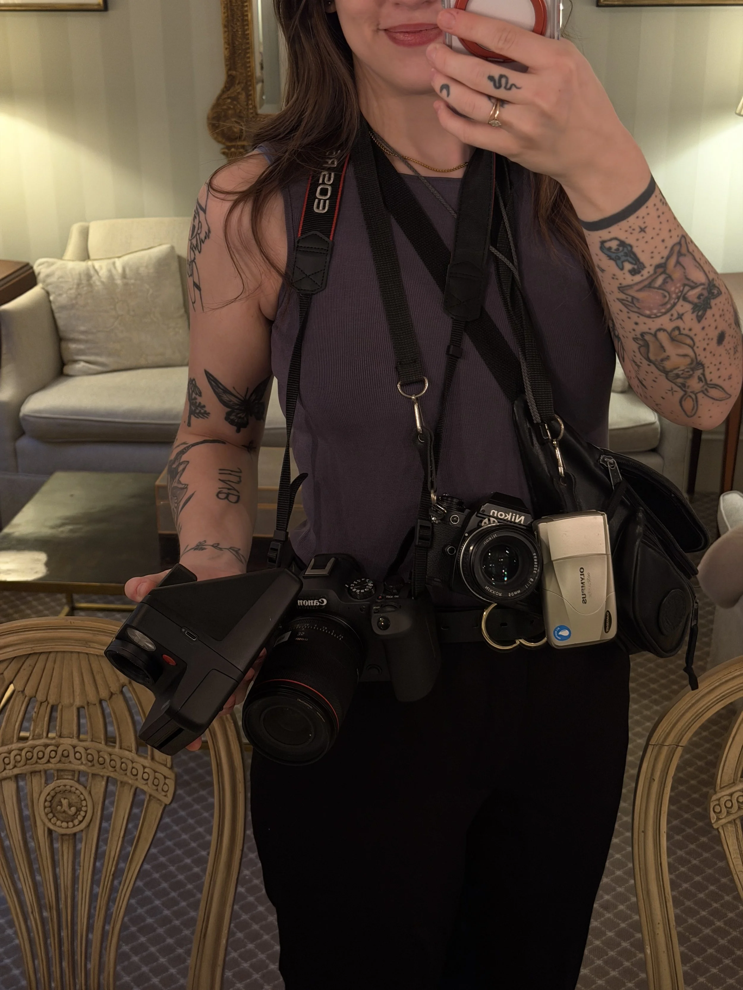 Person taking a selfie, showing the upper body with multiple cameras hanging around their neck and a camera in hand. The person has tattoos on both arms and is wearing a sleeveless top, earrings, and rings. The background features a sofa, a coffee ta