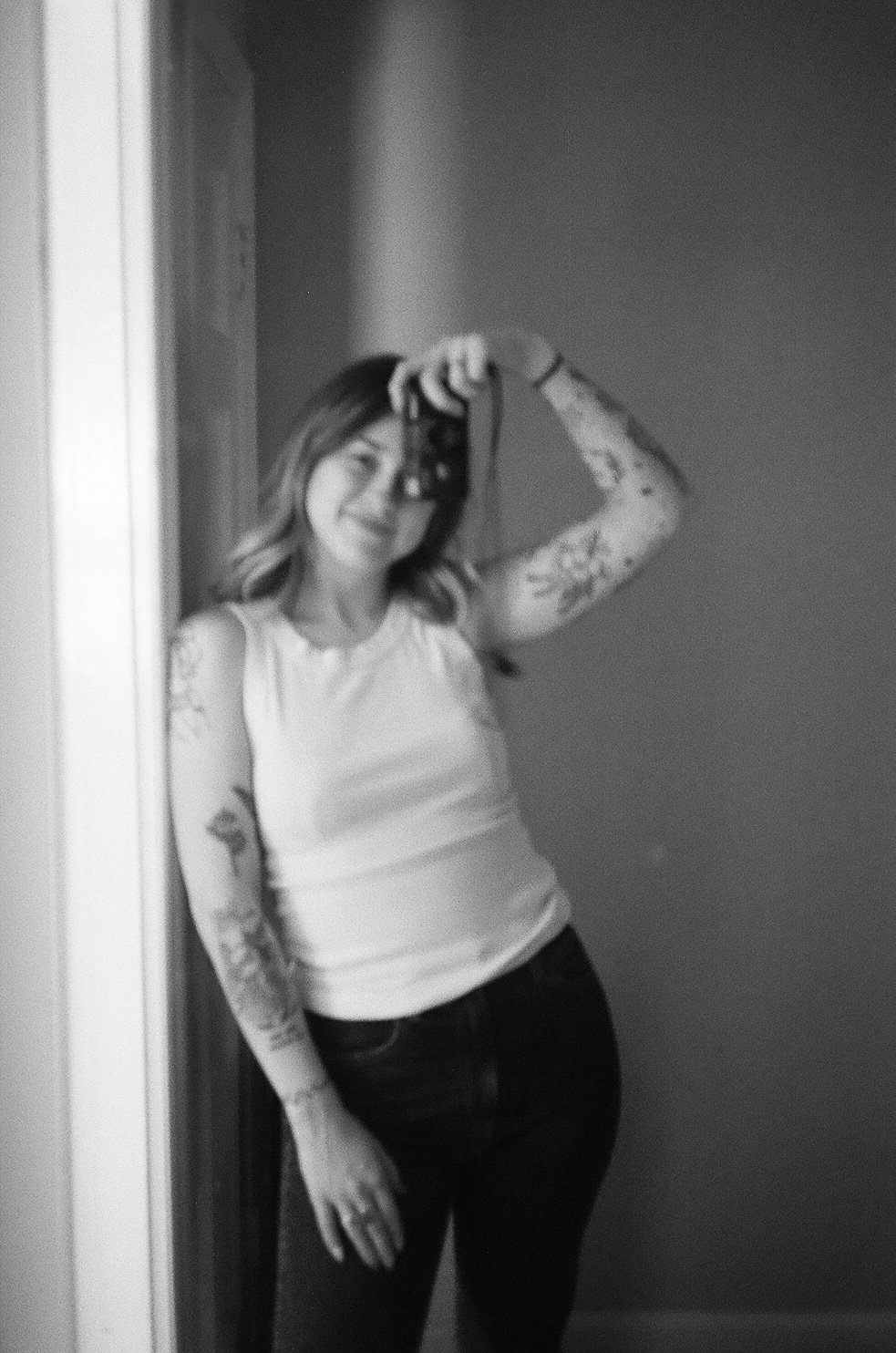 Black and white selfie of a person with tattoos, wearing a tank top and jeans, holding a camera in front of a mirror.