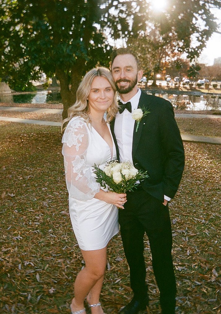 A bride and groom standing outdoors on autumn leaves, smiling, with the bride holding a bouquet of white roses, the groom in a tuxedo with a white rose boutonniere, trees and a pond in the background, and the sun shining through the trees.