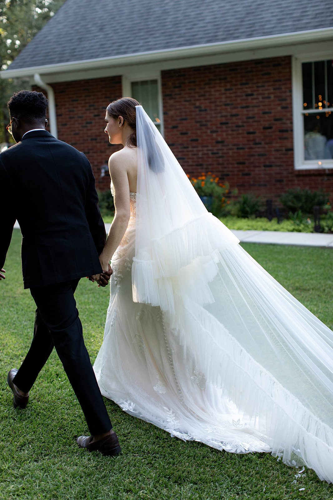 Bride and groom holding hands walking on grass, with the bride wearing a long wedding dress and veil, near a brick building.