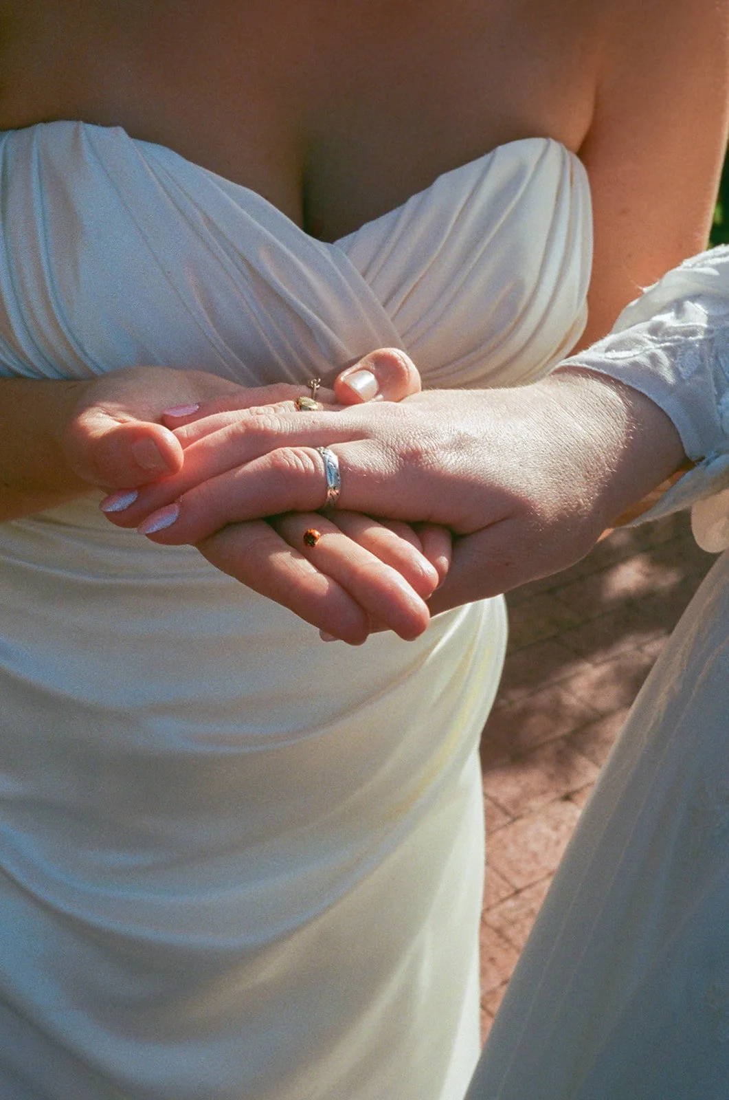Close-up of two people holding hands, with wedding rings on fingers, wearing white clothing, outdoors with natural lighting.