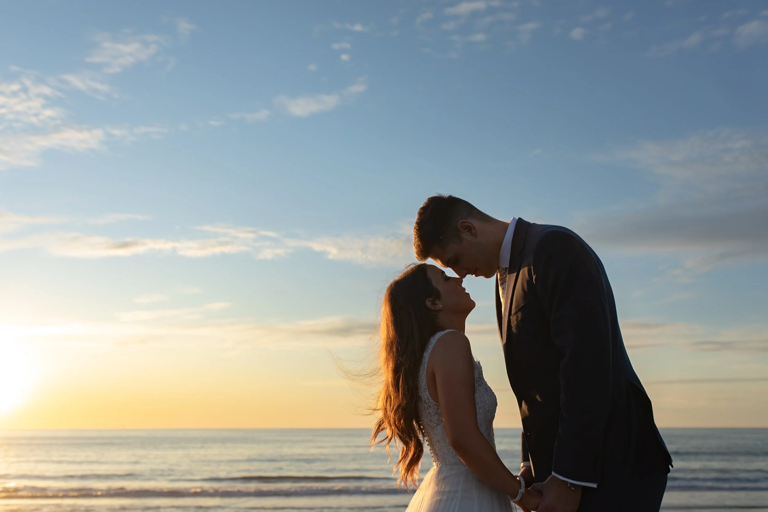 A couple dressed in wedding attire holding hands and touching foreheads on the beach during sunset.