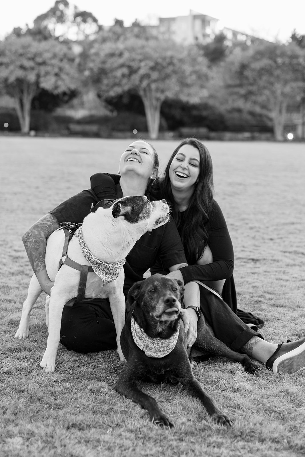 Two people sitting on grass with two dogs, all laughing and smiling. The dogs are wearing bandanas.