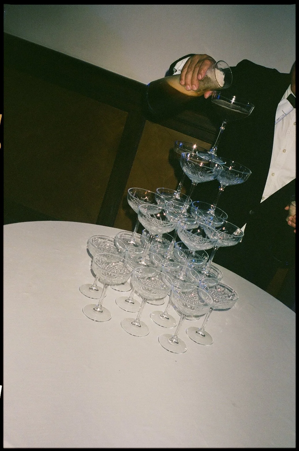 A waiter pouring a drink into the top glass of a pyramid of empty, crystal champagne glasses on a white table.